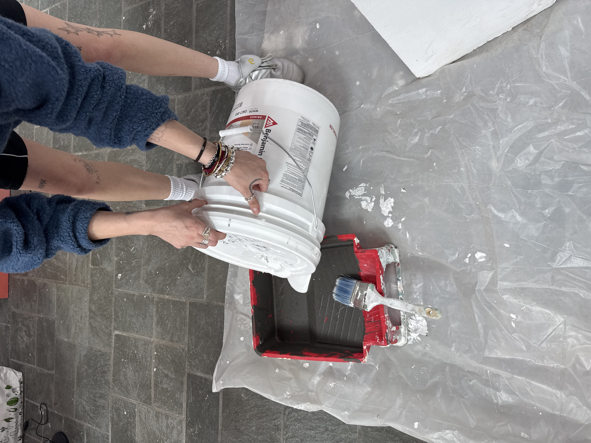 The photograph depicts an individual pouring white paint from a white Benjamin Moore paint bucket into a red paint tray on top of a sheet of clear plastic. The person is wearing a blue sweater and black shorts. Their lower legs and forearms are visible, adorned with tattoos, bracelets, and rings. They are wearing white socks and silver sneakers. A paintbrush is also visible inside the paint tray. The environment includes a gray tile floor, partially covered by the clear plastic sheet. The location is suggested to be Town of Red Hook, United States of America.