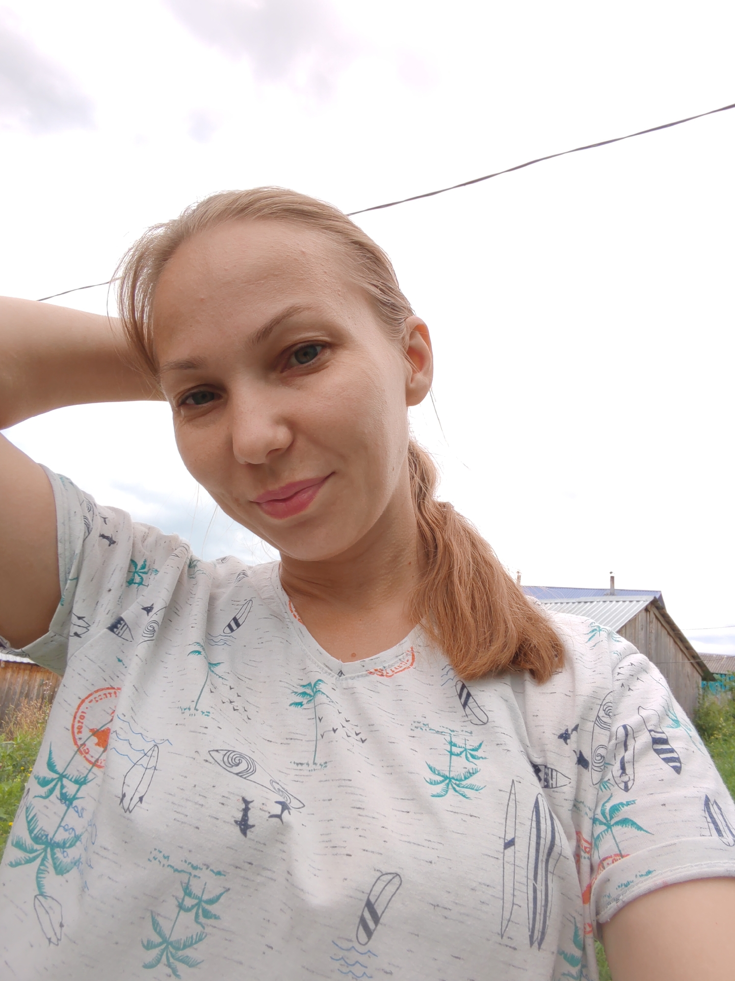 A blonde woman with her hair in a ponytail is taking a selfie outdoors. She is wearing a white t-shirt with a tropical print featuring palm trees and surfboards. Her left arm is raised and bent behind her head. In the background, a wooden building with a metal roof is visible, along with some greenery and a cloudy sky. The scene appears to be in a rural or suburban area in the city of Chukhlem, Russia.
