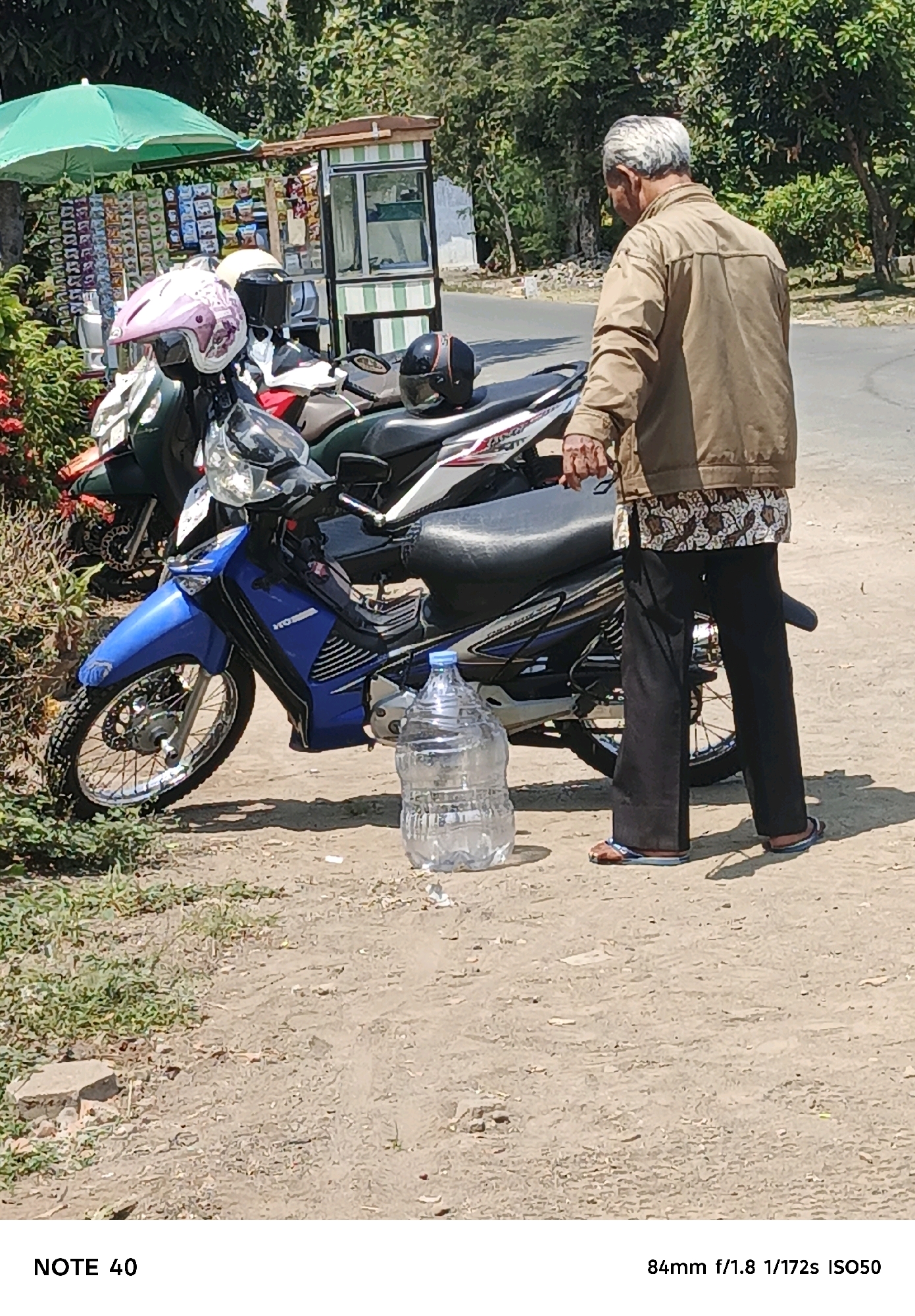 The image depicts an outdoor scene near a roadside stall in Kelurahan Cilacap, Kecamatan Cilacap, Indonesia. In the foreground, a man wearing a tan jacket, a patterned shirt, and black pants with flip-flops stands next to a blue and silver motorcycle. To the left of the motorcycle is a large, clear plastic water jug. Behind the motorcycle, another motorcycle is partially visible, with helmets resting on it. The roadside stall, adorned with green and white stripes, is visible in the background, displaying various items for sale under a green umbrella. The environment appears to be a dusty, unpaved area with sparse vegetation and trees lining the road. The overall impression is one of a typical roadside scene in a local Indonesian setting.