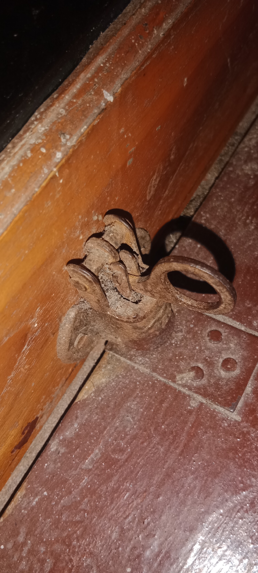 The image shows a close-up view of a rusty metal latch attached to a wooden surface. The latch is old and appears to be made of iron, with intricate detailing and a ring loop. It is fixed to the wood with screws, some of which are also rusted. The surrounding environment suggests an aged and perhaps neglected area, with dust and debris visible on the wood and the floor. The wooden surface appears to be part of a door or furniture, with a reddish-brown hue and visible grain. The floor is also a reddish-brown color and has a slightly textured surface, possibly concrete or painted wood, with a metal plate affixed to it, featuring three circular indentations. The lighting is dim, casting shadows and highlighting the textures of the rusted metal and worn wood. There is no information provided in the image or its context to determine its location in the city of Pelang, Indonesia.