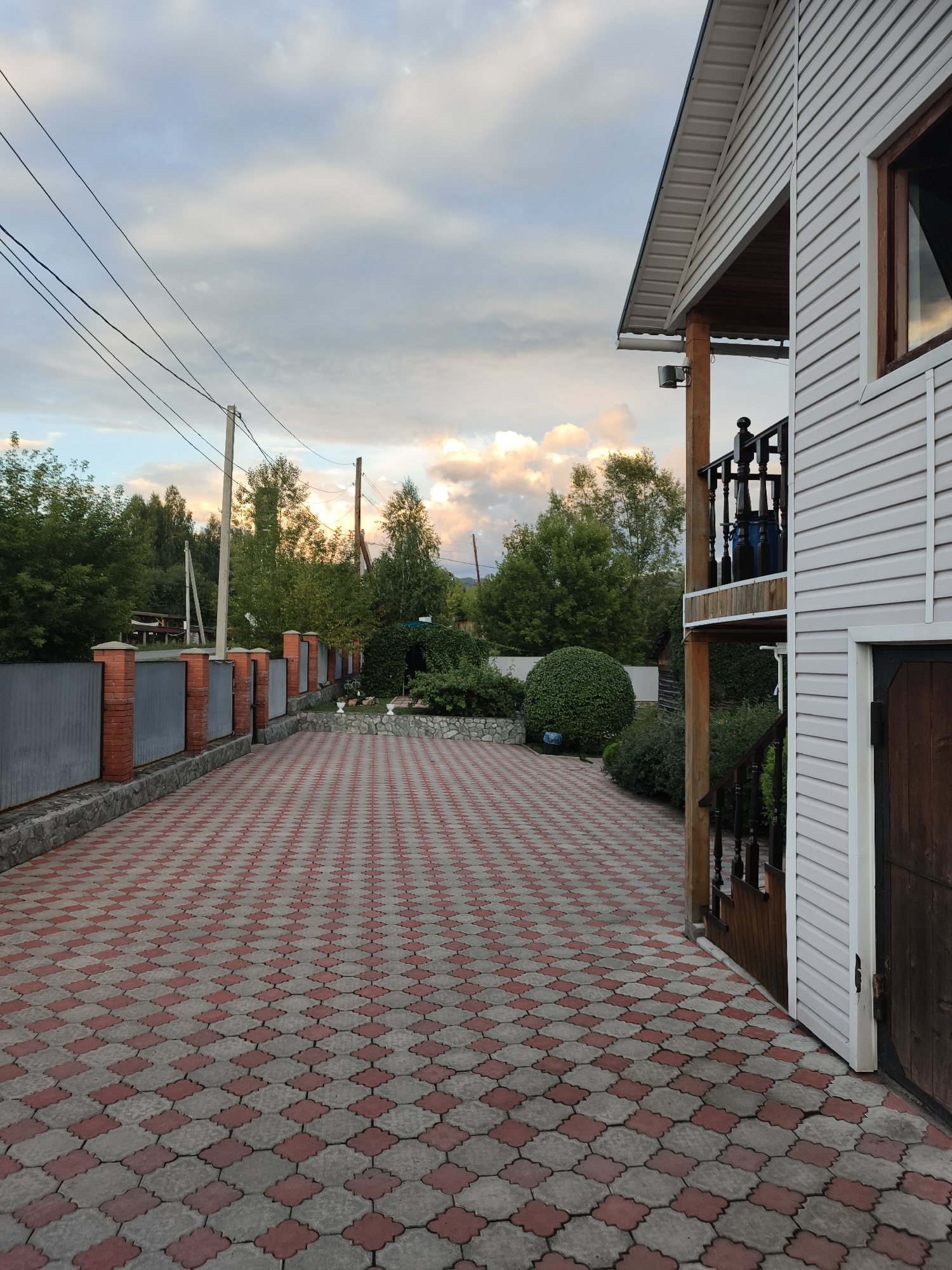The image depicts a paved driveway leading to a white house with a wooden porch and balcony. The driveway is paved with interlocking red and gray pavers arranged in a diamond pattern. To the left of the driveway is a grey metal fence with red brick pillars. Beyond the fence, trees and power lines are visible against a cloudy sky. The house appears to be in a rural or suburban setting, possibly in the city of Katun, Russia, given the architectural style and surrounding vegetation. The time of day appears to be late afternoon or early evening, with the sky showing signs of sunset. There are no people or animals visible in the image.
