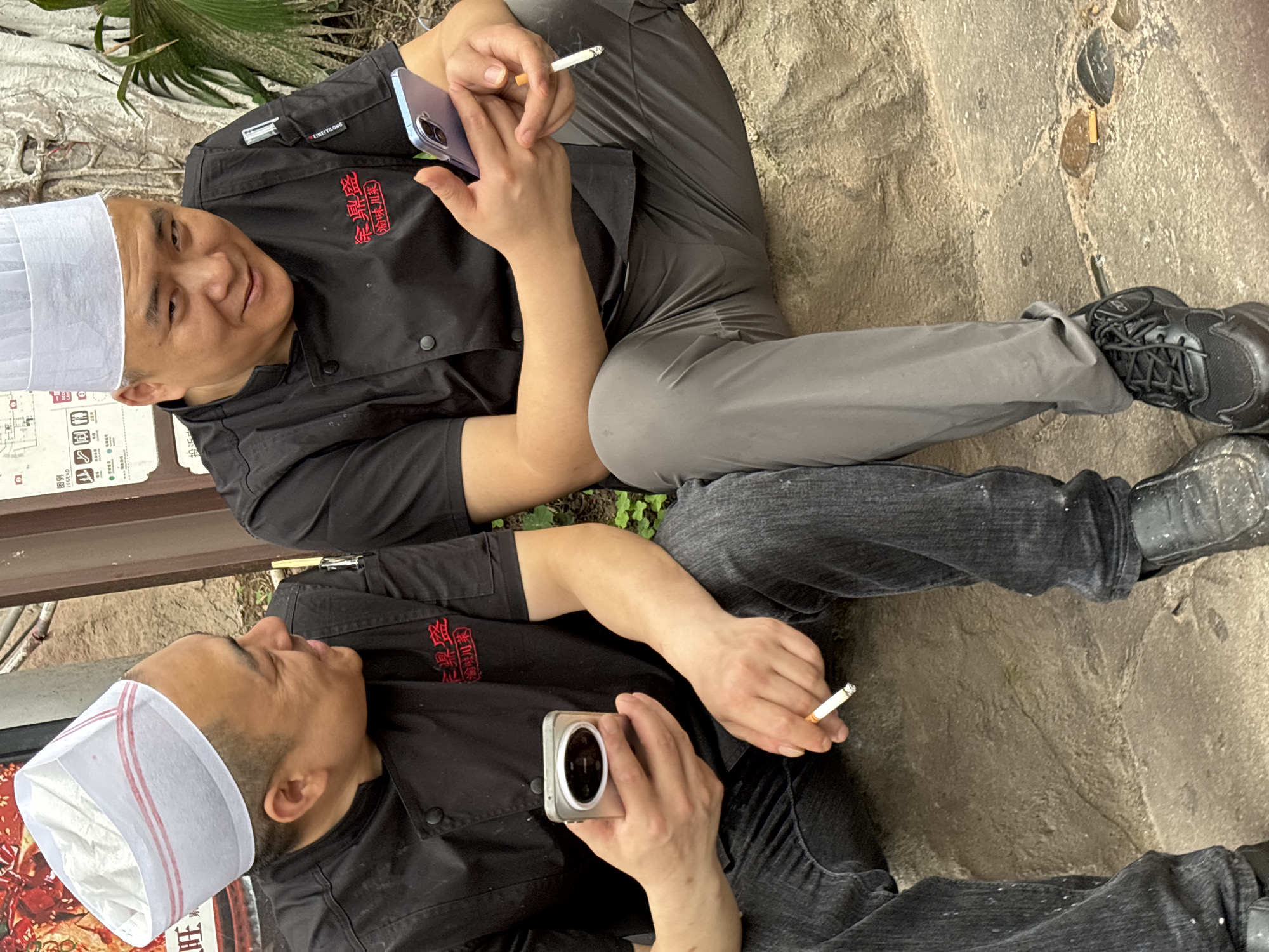 Two male cooks in black chef's coats and white hats sit on the ground. One holds a blue smartphone and a lit cigarette. The other holds a silver compact camera and a lit cigarette. Their clothing includes grey sweatpants and black jeans. The location is indicated by Chinese text on the coats, "徐鼎盛(渝味川菜)" and "EIMEI YILONG", and nearby signage with "图例 LEGEND". The location is Yuzhong District, China.