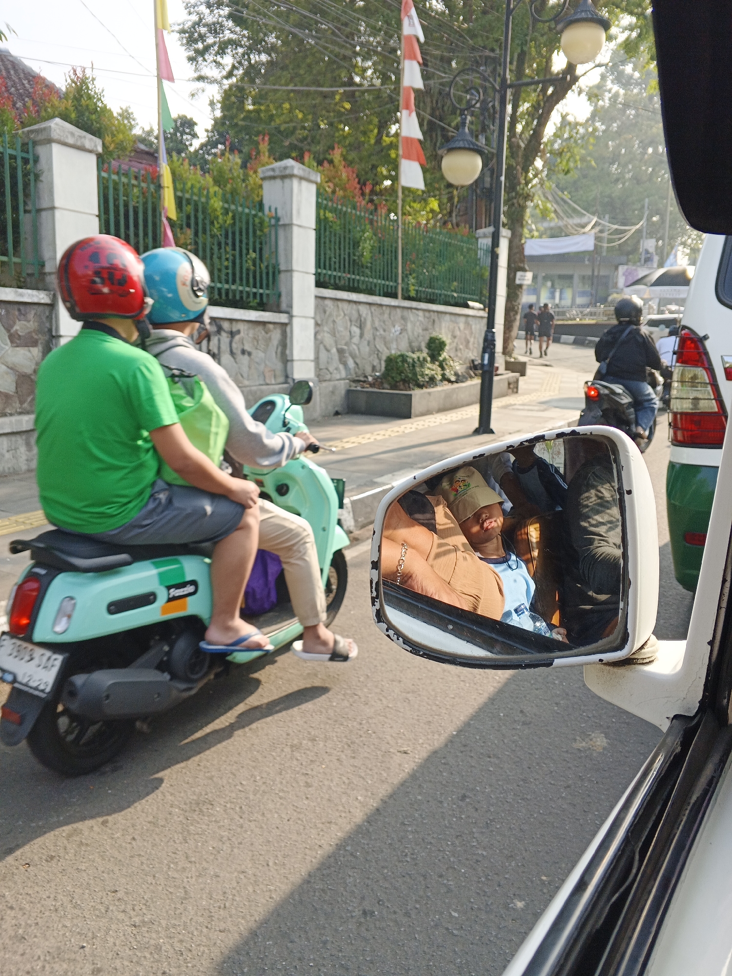 In Sukabumi, Indonesia, two individuals are riding a light blue scooter on a street bathed in sunlight. The person in front wears a green shirt and a red helmet, while the passenger behind them has a light gray hoodie and a blue helmet. A side-view mirror reflects a child wearing a beige hat and a light blue shirt, asleep while sitting next to an adult. The scooter displays a license plate that reads "F 3909 SAF". In the background, a white vehicle with a green stripe is partially visible, suggesting it is also on the road. The environment includes a sidewalk, a stone wall, green fences, and trees.