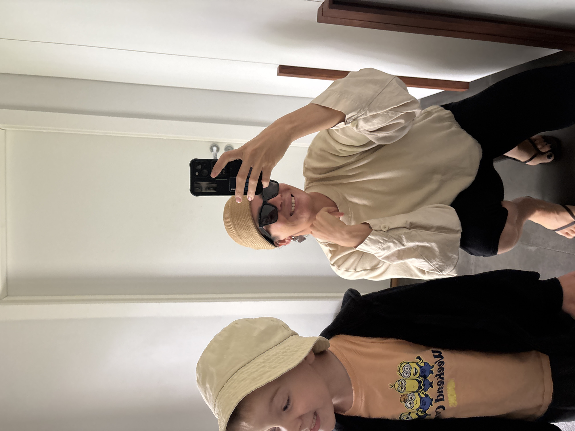 In a brightly lit indoor setting, a woman and a young boy are captured in a mirror selfie. The woman, on the right, is wearing a tan beanie, sunglasses, and a light-colored, loose-fitting shirt with voluminous sleeves. She is holding a smartphone, taking the picture, and has her hand on her chin, smiling. The boy, on the left, is wearing a light tan bucket hat, a light orange t-shirt with a Minions graphic, and a black zip-up jacket. He is looking to his left and smiling. They appear to be in a domestic environment, possibly a hallway or room with a white door and wall visible. The flooring is dark. The image does not provide any specific details to indicate the location is the city of Chaikovsky, Russia.