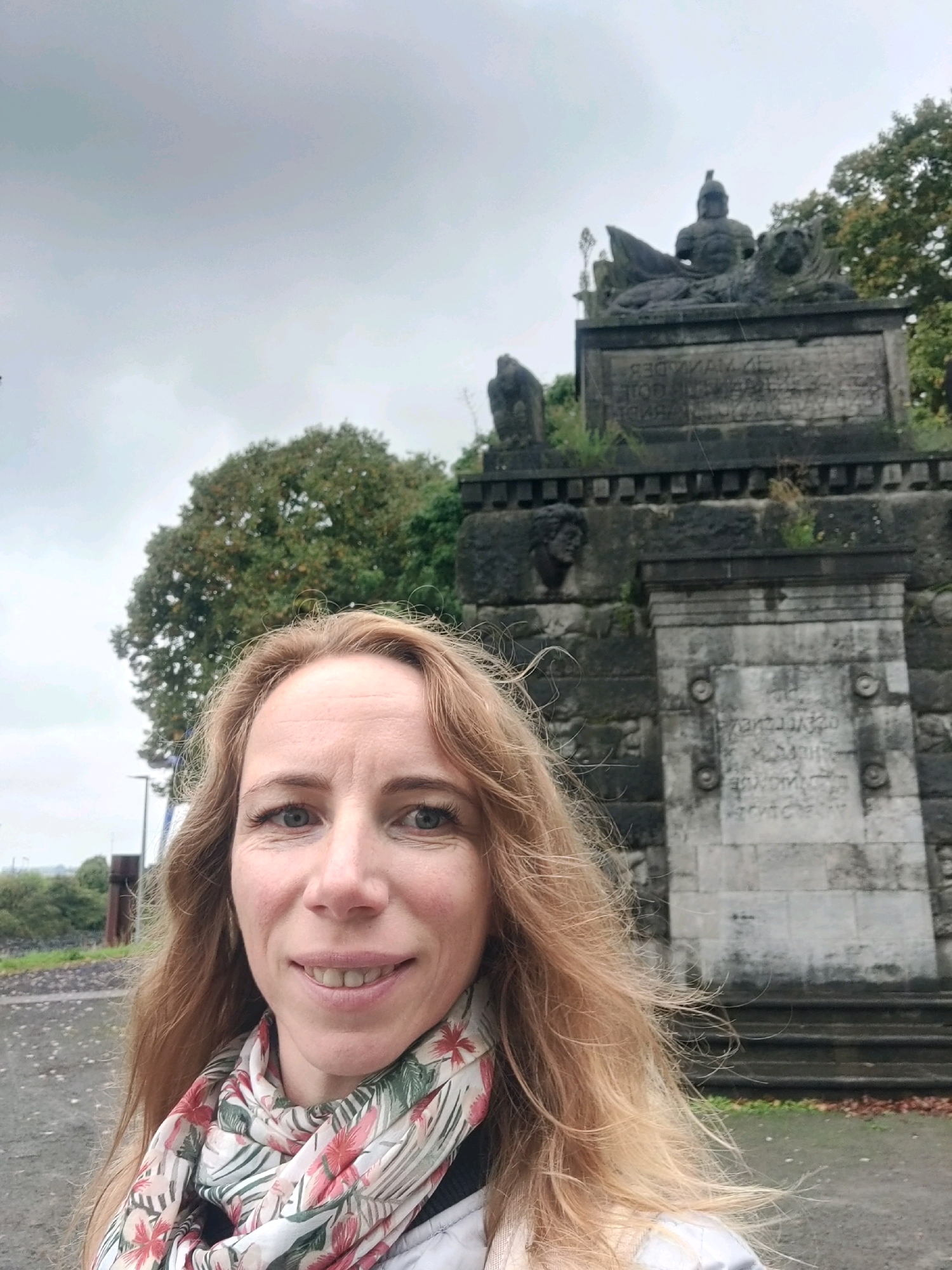 A blonde woman smiles at the camera in a selfie taken in front of a stone monument. She is wearing a scarf with a floral pattern of pink and green flowers. The monument behind her appears to be made of large stone blocks, with carvings and inscriptions. One carving is of a soldier with a helmet and shield, lying on a lion. Other carvings include what appear to be faces and a bird of prey. The sky is overcast and grey, and there are trees with green and autumn foliage visible behind the monument. The ground in front of the monument is paved with asphalt and gravel. The location is identified as Emmerich on the Rhine, Germany.