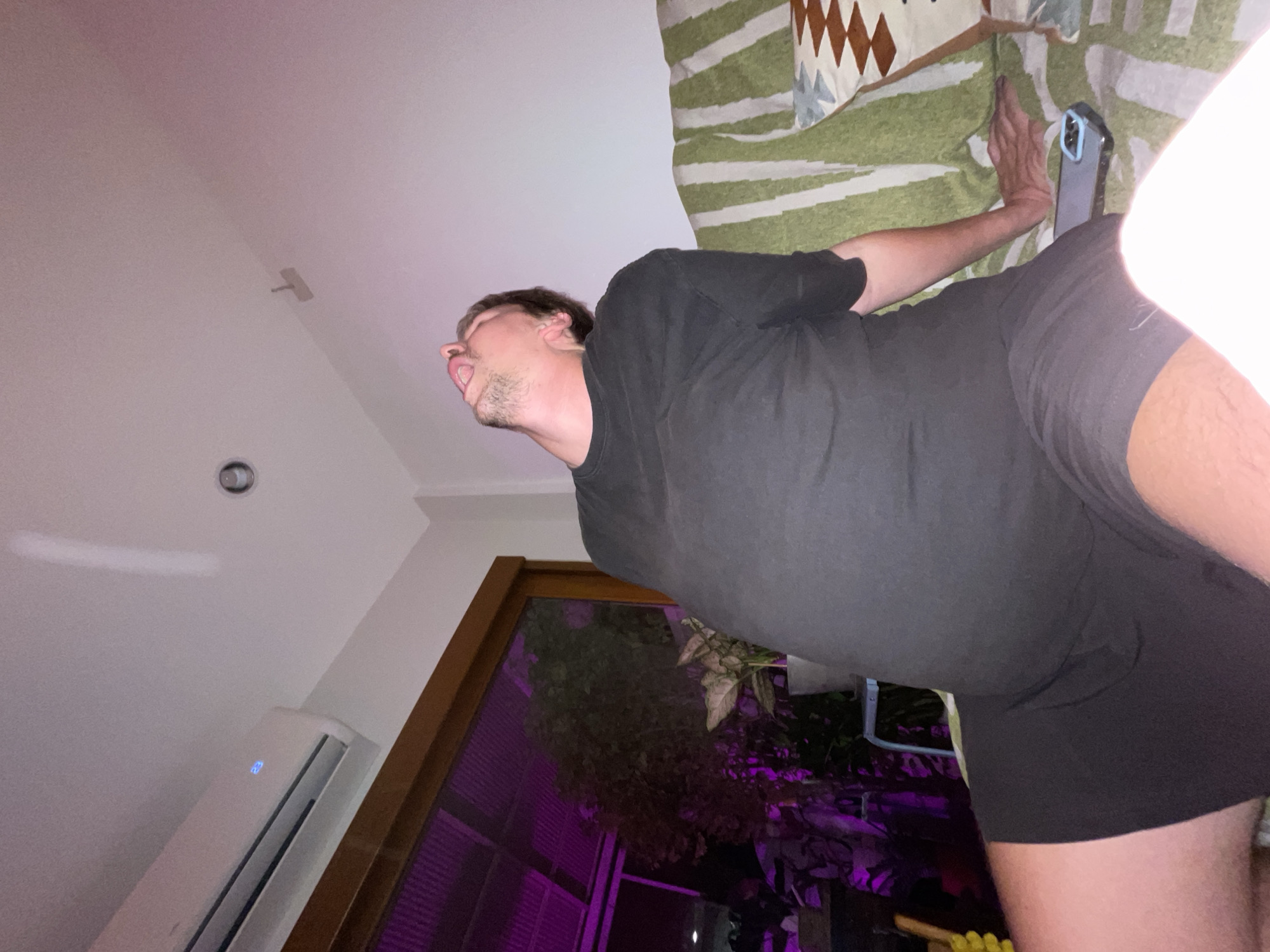 A man is captured lying down indoors, viewed from a low angle. He is oriented horizontally across the frame, wearing a black t-shirt and black shorts. His head is tilted upwards with his mouth slightly open, revealing some facial stubble. His left arm is bent behind him, and his hand rests near a smartphone in a light blue case, which lies on a patterned fabric featuring green and white stripes.
The background shows a white ceiling with a recessed light and a small vent. An air conditioning unit with a blue digital display is mounted on a nearby wall. A large window or glass door with a dark wooden frame is visible, looking out onto outdoor foliage that is distinctly illuminated with a purple light, suggesting artificial lighting at night. The man's bare right leg is partially visible in the foreground. The setting is an indoor space in Chang Phueak, Thailand, likely in the evening or at night. No readable text is visible.
