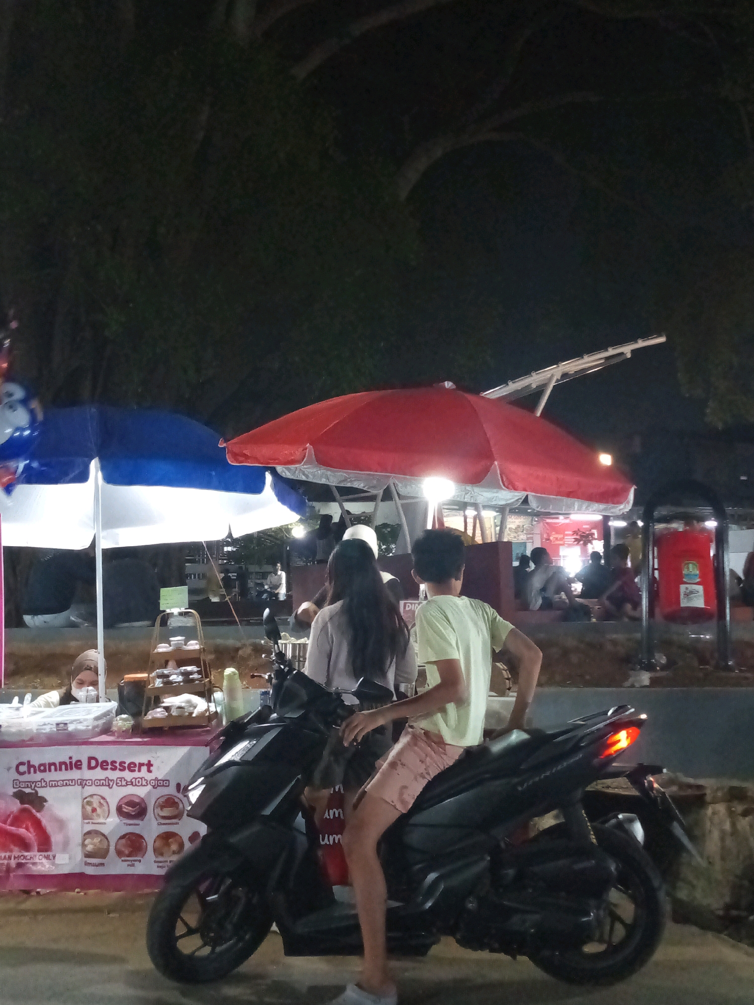 In Bekasi, Indonesia, at night, a black motorbike is parked in front of a dessert stall. Two people are on the motorbike, one in a light green t-shirt and pink shorts, the other with long dark hair wearing a light brown jacket. A blue and white umbrella and a red umbrella are set up over the stall, which displays various desserts and has a banner that reads "Channie Dessert". Other people are visible in the background, some sitting at tables. The scene is illuminated by streetlights and the stall's lighting.