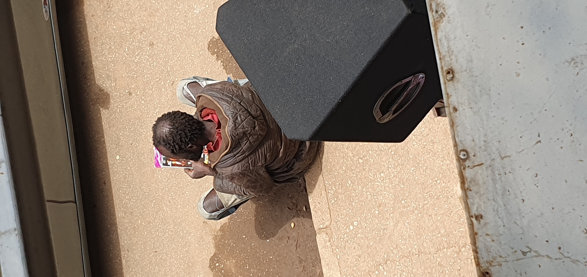 A person is sitting on a raised step, facing away from the camera and looking at a tablet. The person is wearing a brown quilted jacket, a red shirt, and ripped light-colored pants. The person has short, curly, dark hair and appears to be male. The person is sitting on concrete steps. To the right of the person, a large black speaker is partially visible. The ground is a mixture of dirt and small stones. In the background, a grey wall or structure is visible. The scene appears to be outdoors in an urban environment, possibly in Bukuru, Nigeria.