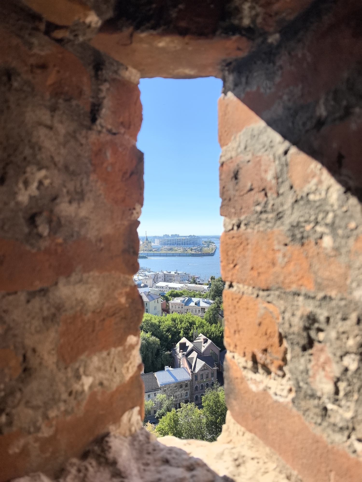 The image, captured through a gap in a brick structure, offers a view of the city of Nizhny Novgorod, Russia. In the foreground, the rough texture of weathered red brick walls is visible, with mortar filling the gaps between the bricks. The aperture frames a distant cityscape dominated by a wide river, likely the Volga River, with a large white building complex situated on its far bank under a clear blue sky. Closer to the viewer, a cluster of traditional European-style buildings with pitched roofs and varied architectural details are nestled among lush green trees. A prominent yellow-domed church stands out amidst the greenery, and further down the river, there are indications of boat traffic. The sunlight casts shadows across the brickwork in the foreground, suggesting it is a bright, sunny day.