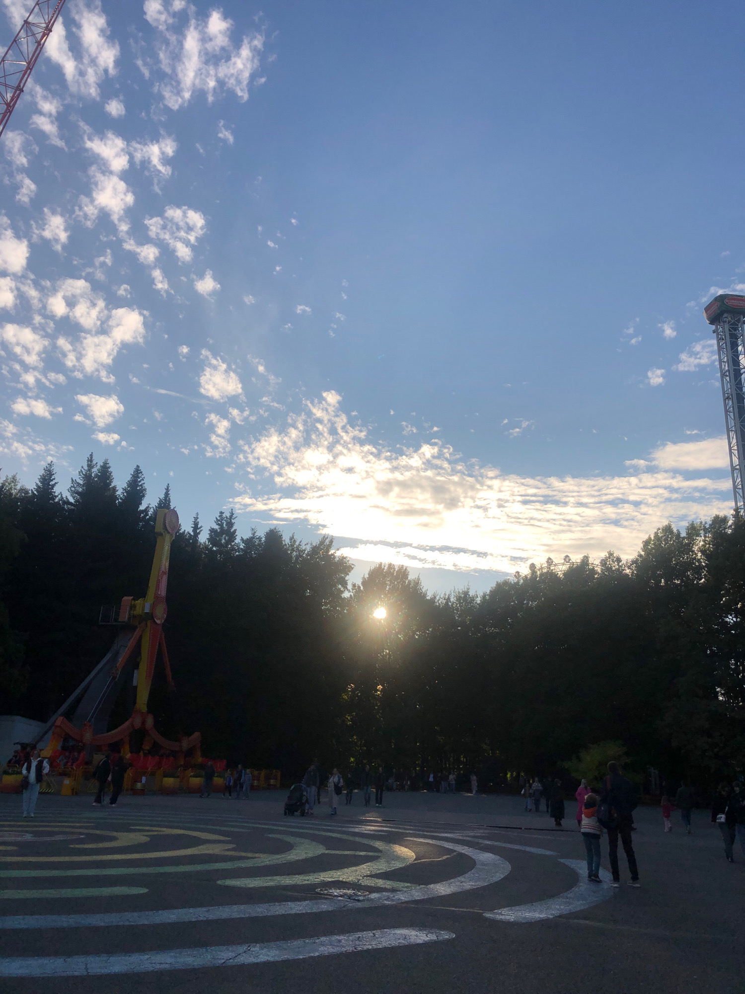 This is a horizontal, eye-level, medium-shot photograph of an amusement park. In the foreground, a paved area has a colorful, maze-like design painted on it. Several people are walking in the paved area, some are silhouetted by the bright sun. In the midground, a large amusement park ride with yellow and red arms is visible. Behind the ride and a line of dark green trees, the sun is setting, casting a bright glare in the center of the image. On the right side of the image, a tall tower ride with a red top is visible against a light blue sky with scattered white clouds. The atmosphere is that of an amusement park at dusk. The location is likely not Saint Petersburg, Russia.