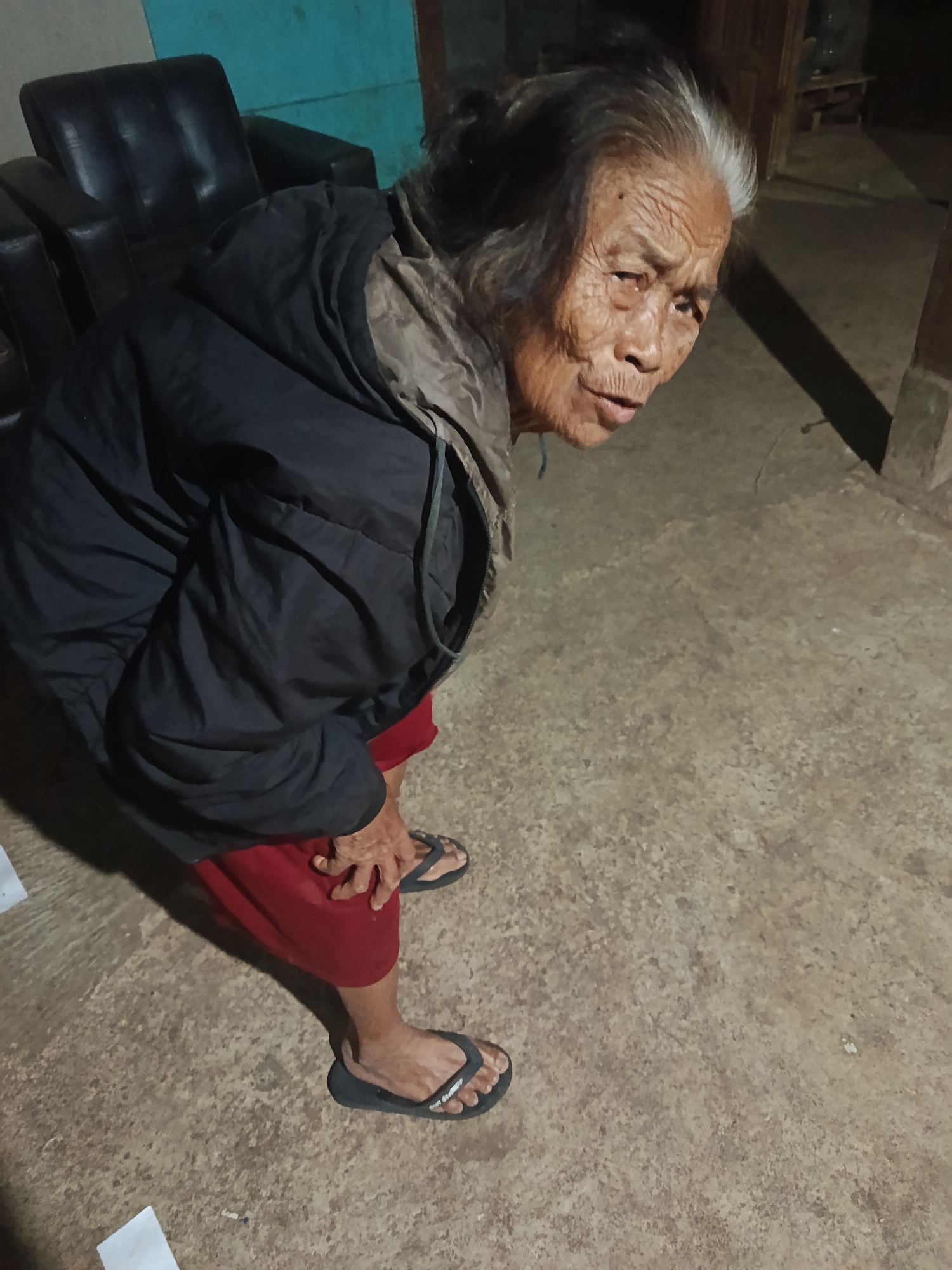 An elderly woman with graying hair and wrinkled skin is crouching on a concrete floor in what appears to be a dimly lit room. She is wearing a dark jacket over a red garment and black flip-flops. Her left hand is resting on her knee, and her body is angled towards the right side of the frame. In the background, a black leather armchair is visible, and the walls are painted a teal color. The scene suggests an indoor setting, possibly a residence in Karangmojo, Indonesia.