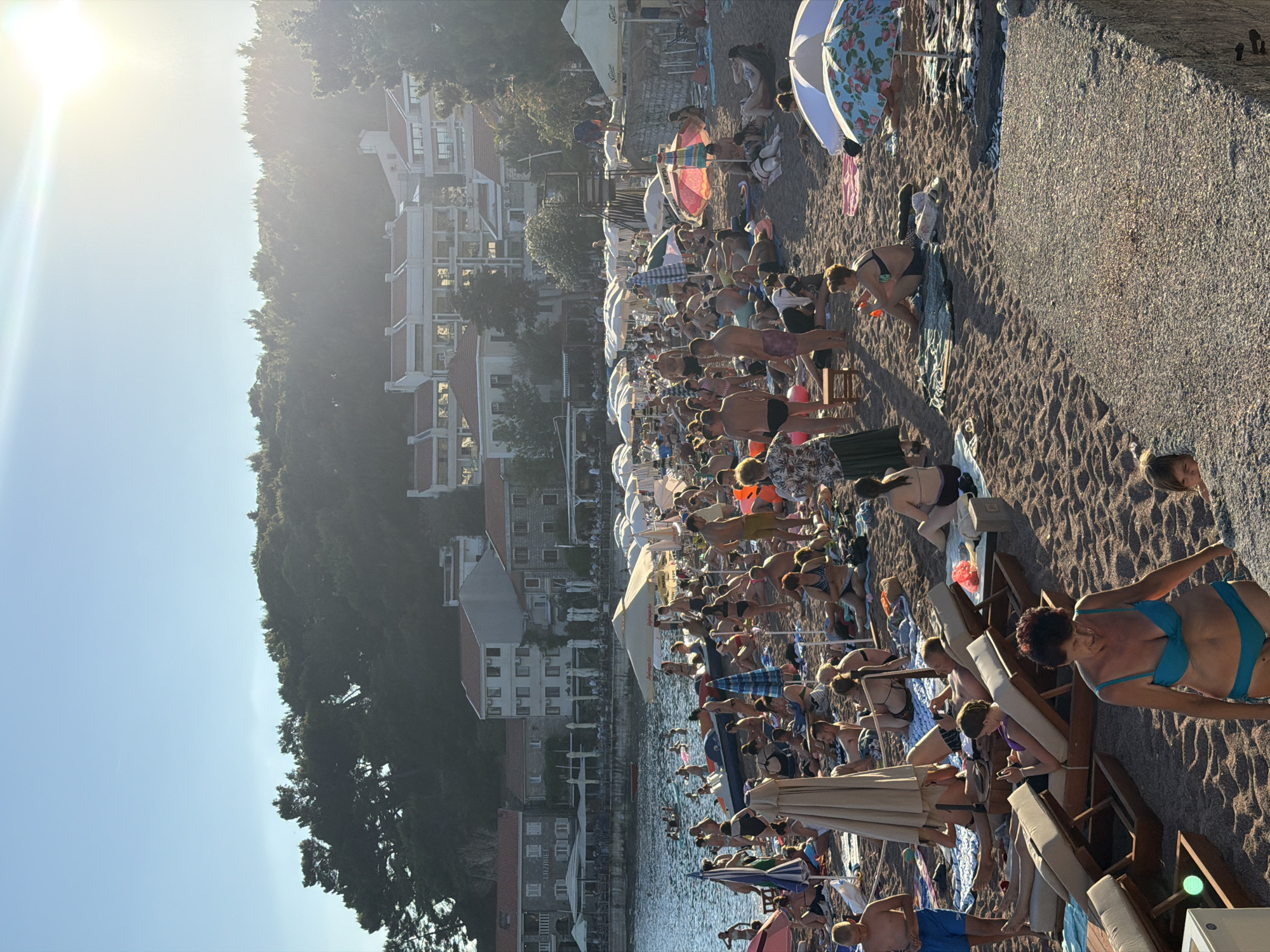 The image depicts a crowded beach scene in Petrovac centar, Montenegro, bathed in the warm glow of a setting sun. The sandy beach is densely populated with numerous individuals of various ages, many of whom are dressed in swimwear and lounging on towels or beach chairs. Some people are actively engaged in swimming or wading in the clear blue water, while others are seen walking or interacting on the shore. A variety of beach umbrellas, some colorful and others white, are spread across the sand, providing shade to beachgoers. In the background, a line of buildings, likely hotels or apartments with red-tiled roofs, are visible, overlooking the beach and the sea. Lush green trees and vegetation cover the hillsides behind the buildings. The overall atmosphere is one of a bustling summer vacation spot, with people enjoying leisure activities by the coast.