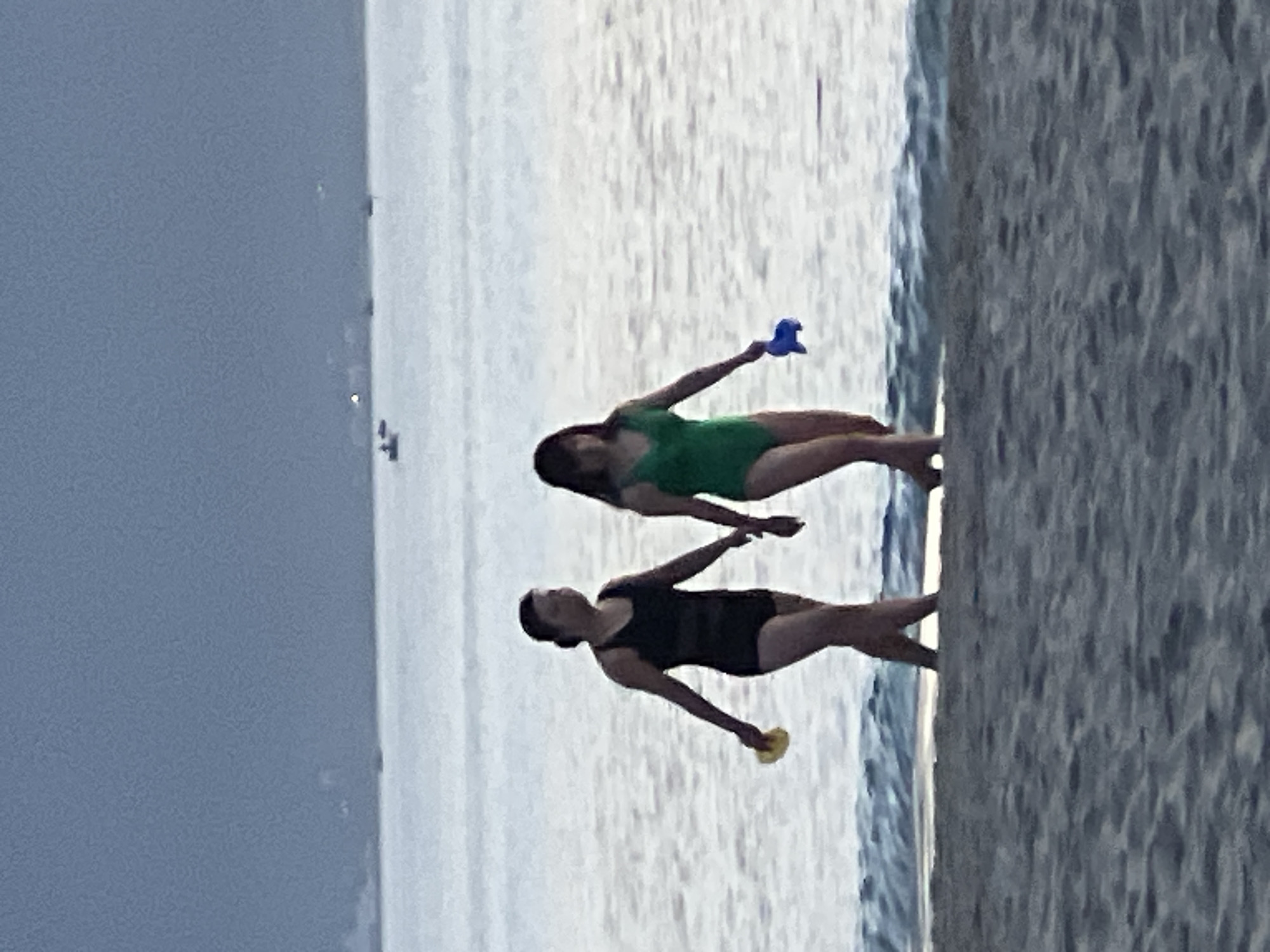 Two individuals are walking on a sandy beach in Đà Nẵng, Vietnam, near the ocean. The person on the left is wearing a black one-piece swimsuit and holding a yellow object in their left hand. The person on the right is wearing a green one-piece swimsuit and holding a blue object in their right hand. The ocean is calm with small waves lapping at the shore. In the distance, several small boats and possibly a distant shoreline with lights are visible under a hazy sky. The sand in the foreground is textured with small pebbles and footprints. The lighting suggests it is either early morning or late evening.