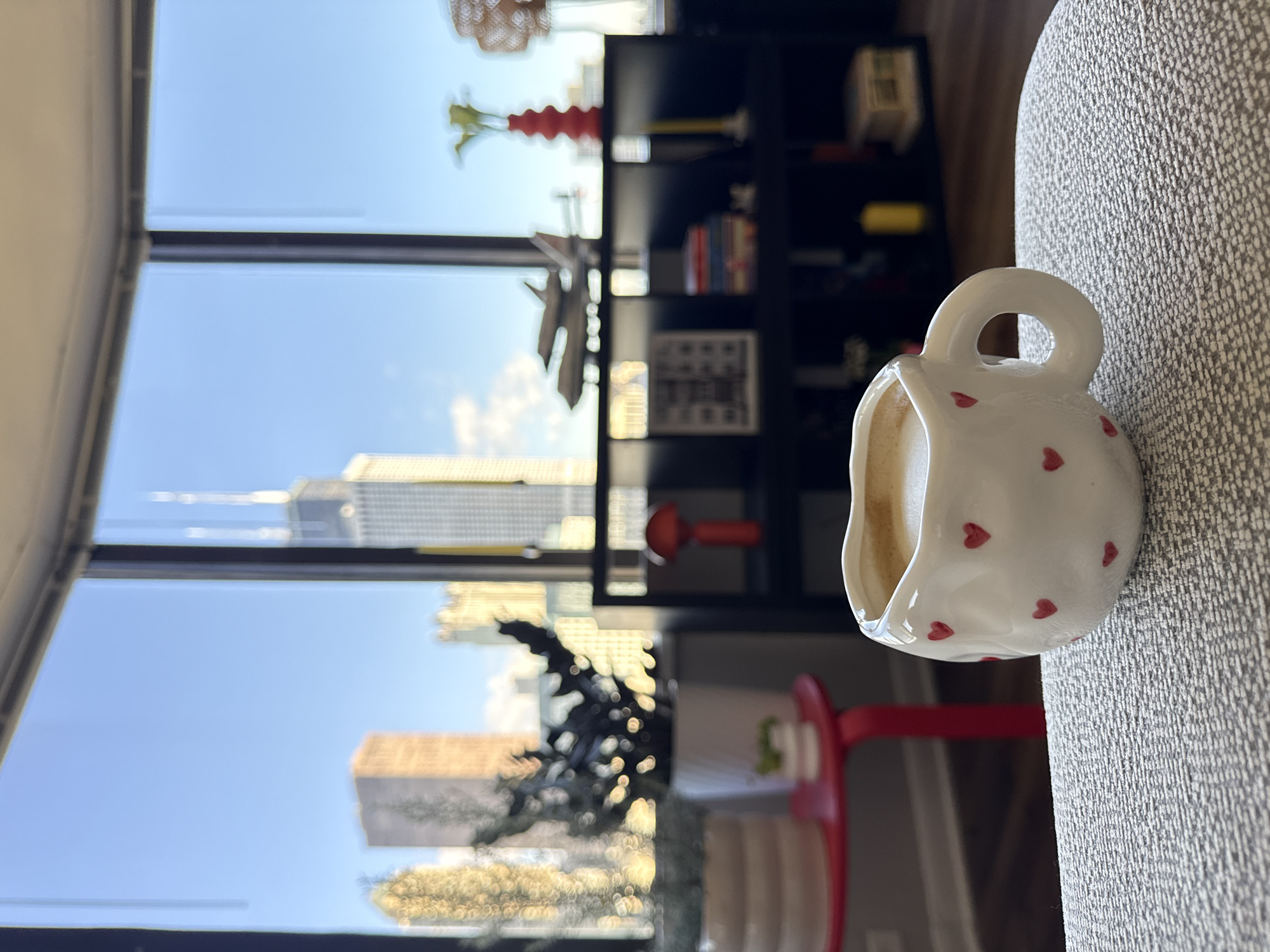 The media file shows a white ceramic mug with red hearts on it, filled with a light-colored beverage, possibly coffee. The mug is positioned on a textured, light-colored surface, which appears to be upholstery, possibly a sofa or chair. In the background, through a large window, a cityscape is visible under a clear blue sky, suggesting daytime. Prominent buildings, including skyscrapers, are seen, hinting at an urban environment. Reflections in the window add to the scene's depth. The setting appears to be indoors, likely an apartment or office with a view of Chicago. There are shelves filled with books and decorative items, and a glimpse of red furniture. The overall impression is a moment of quiet contemplation or relaxation. No people are visible. There is no visible text.