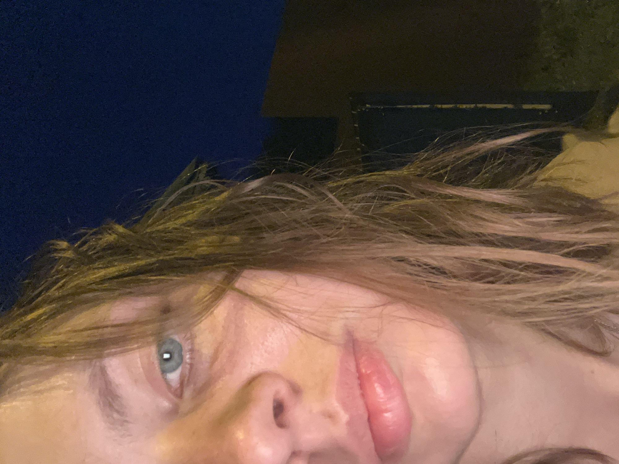 The image is a close-up, medium shot of a young woman's face and upper body, taken at night. Only the left side of her face is visible, including her left eye, nose, and lips. Her eye is a light blue or green, and her lips are slightly parted and appear to be glossy. Her hair is long and brown, with some strands falling across her face. She is wearing a light-colored garment that is partially visible. The background is dark, with a deep blue sky and the outline of a building. There is also some foliage visible in the lower right portion of the image. There is no specific information in the image that would indicate it was taken in Nizhny Tagil, Russia.