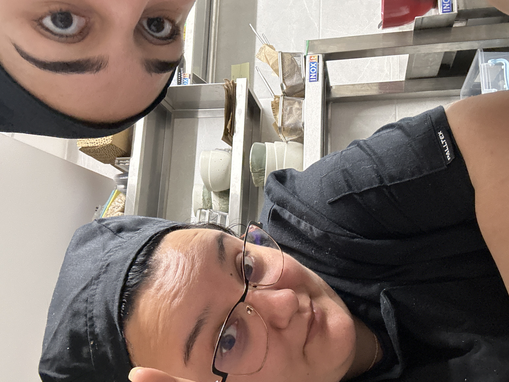 Two individuals are captured in a close-up, tilted selfie. Both appear to be wearing black head coverings. One individual has thick eyebrows and is positioned closer to the top edge of the frame, with only their eyes and a portion of their forehead visible. The other person is wearing glasses and looking directly at the camera. They have fair skin and are wearing a black uniform.
The background includes metallic shelving, containers, and what appears to be a logo that reads "INOX" on a blue label. There are also paper-lined containers stacked on the shelves. The brand name "PALLTEX" is visible on the upper right side of the frame.
There is insufficient information to identify the location of Burgas, Bulgaria.