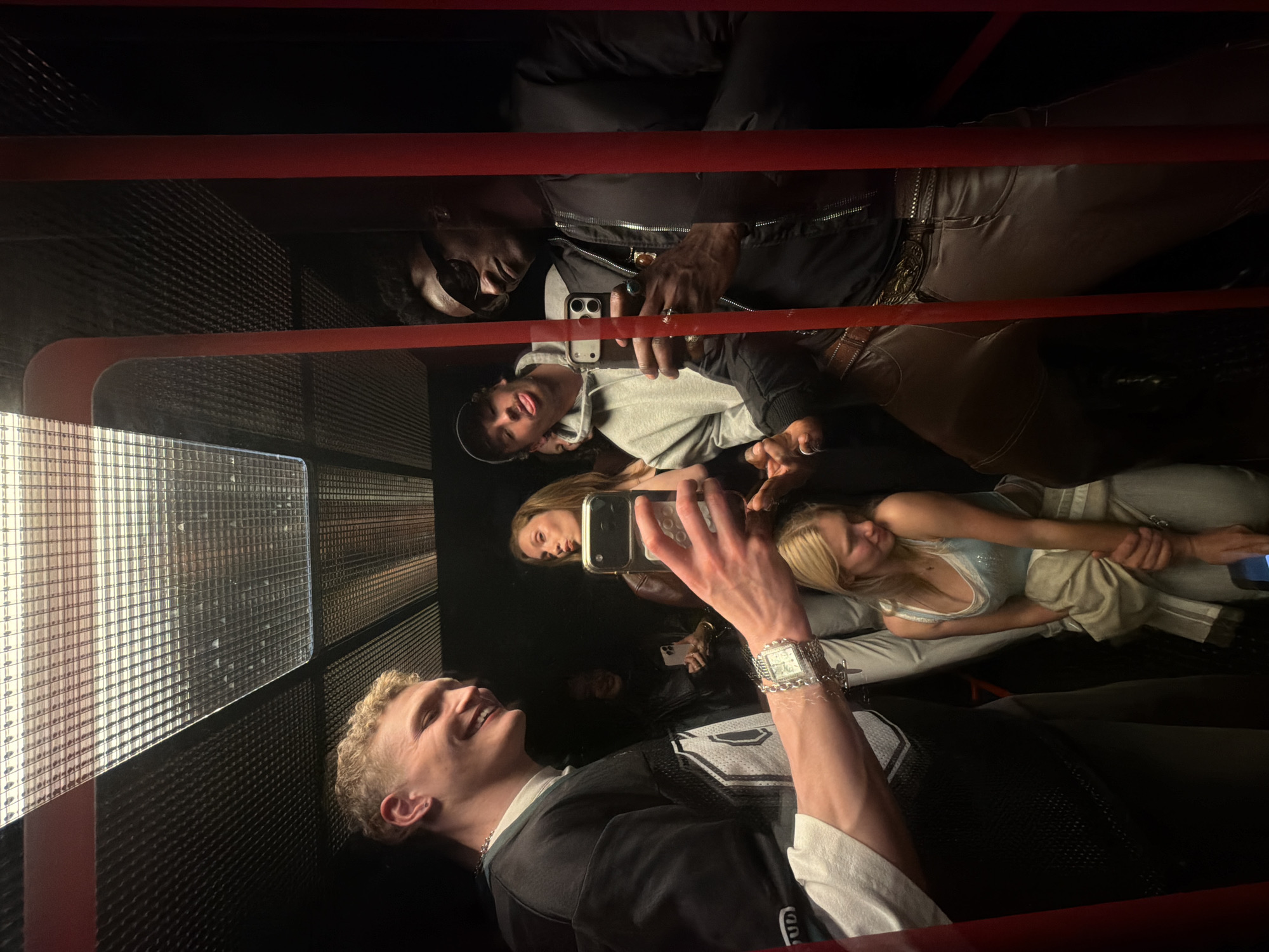The image shows a group of people in what appears to be an elevator or small room with mirrored walls. There are approximately six individuals visible, with a primary focus on the person in the foreground who is smiling and holding a phone, possibly taking a selfie or video. The room has a grid-like wall on one side, allowing light to enter. The elevator shaft is visible, with some red bars. The people seem to be in a lively, social mood, likely traveling between floors of a building. Given the style and location, it is not possible to be precise, but the people are likely traveling in a London skyscraper.