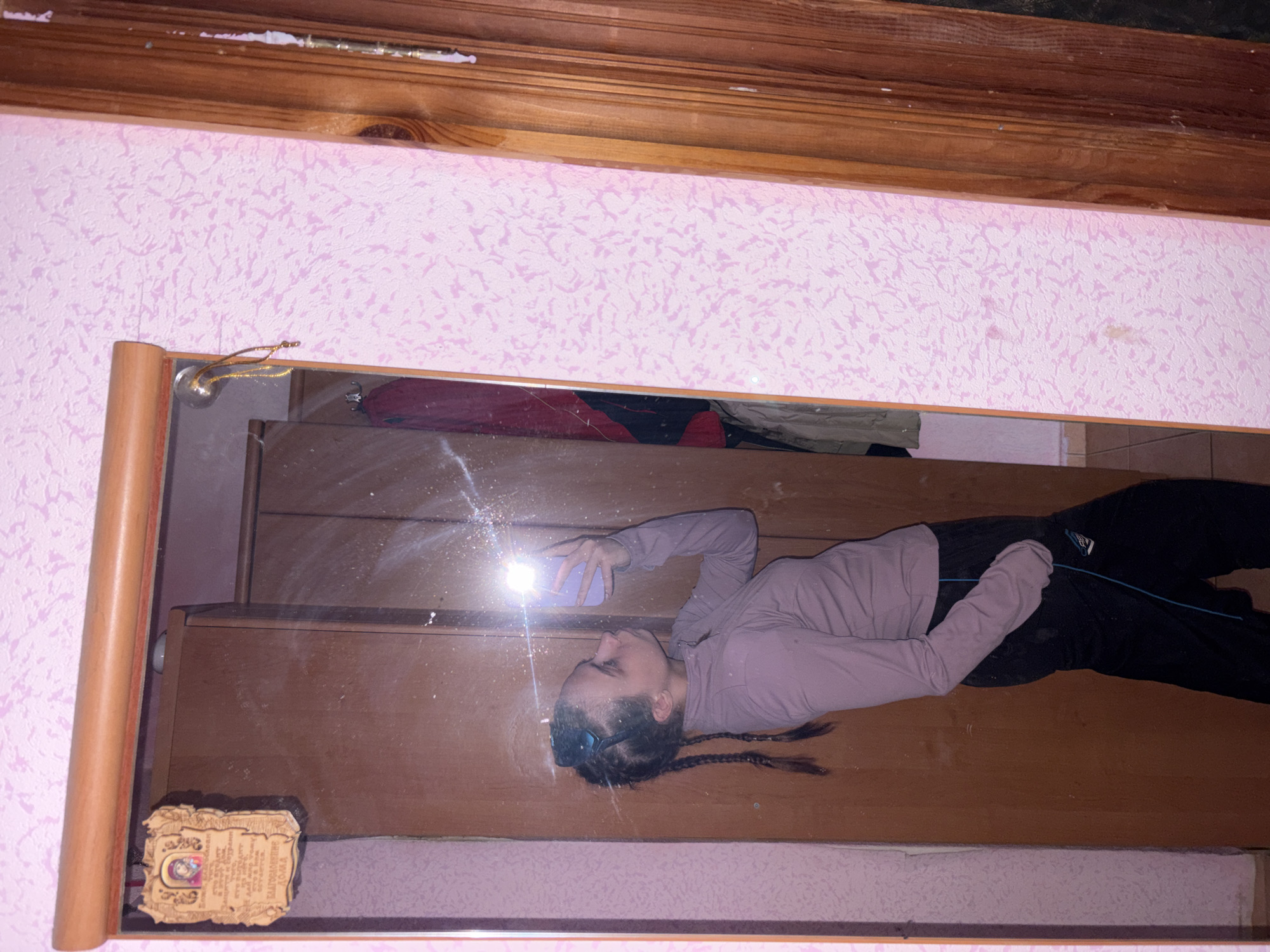 The image, originally oriented sideways, depicts a person taking a mirror selfie indoors, likely in Прикамский, Russia. A young individual, reflected in a large, wooden-framed mirror, is the central subject. They are dressed in a light mauve long-sleeved top and dark pants, with dark hair styled in two braids, and sunglasses resting on their head. The person is holding a light-colored smartphone, aimed towards themselves, with the camera flash brightly illuminating their face and causing a strong glare in the reflection.
The visible interior includes pink textured wallpaper with a subtle pattern, bordering the mirror above and below. Wooden trim runs along the top and bottom edges of the wall. Within the mirror's reflection, dark wooden furniture, possibly a dresser or cabinet, is visible in the background. A small, ornate plaque or icon is affixed to the mirror frame in the bottom left corner. This plaque features Cyrillic text, clearly displaying "БЛАГОСЛОВЕНЬЕ ДОМА" (Blessing of the Home), with other smaller, illegible script. The setting appears to be a casual residential space, and the use of flash suggests either low ambient light or a deliberate photographic choice.