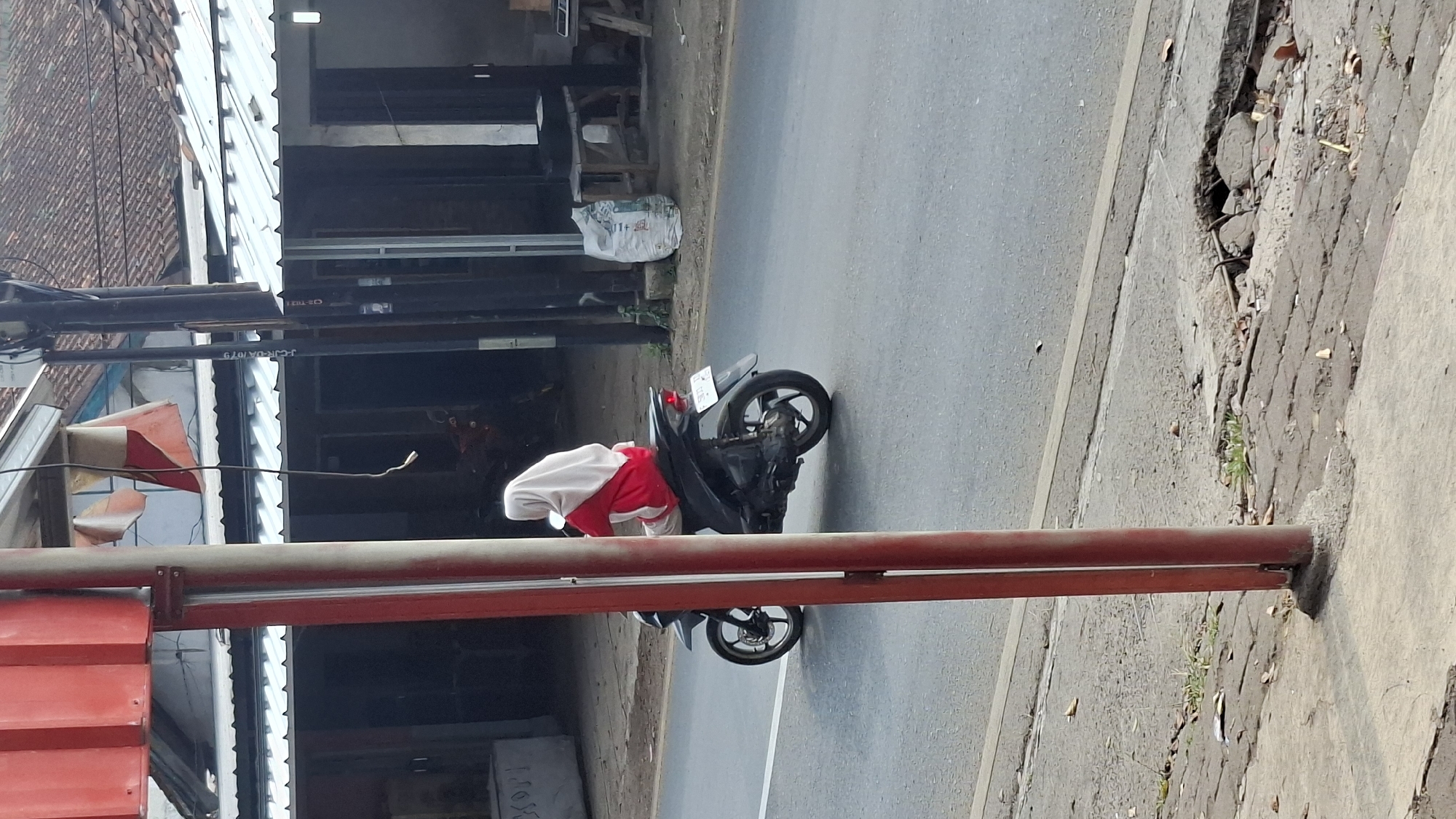 A side-view, medium shot shows a person wearing a white hijab and a red top riding a black motorcycle on a paved road in Cilaku, Indonesia. The motorcycle is moving away from the camera towards the right side of the frame. The road has a white line marking the lane. In the foreground, two vertical red poles are visible, with one slightly out of focus. Behind the motorcycle, there are buildings with tiled roofs, power poles, and a white sack leaning against a post. The environment appears to be a street scene with some wear and tear on the pavement in the foreground.