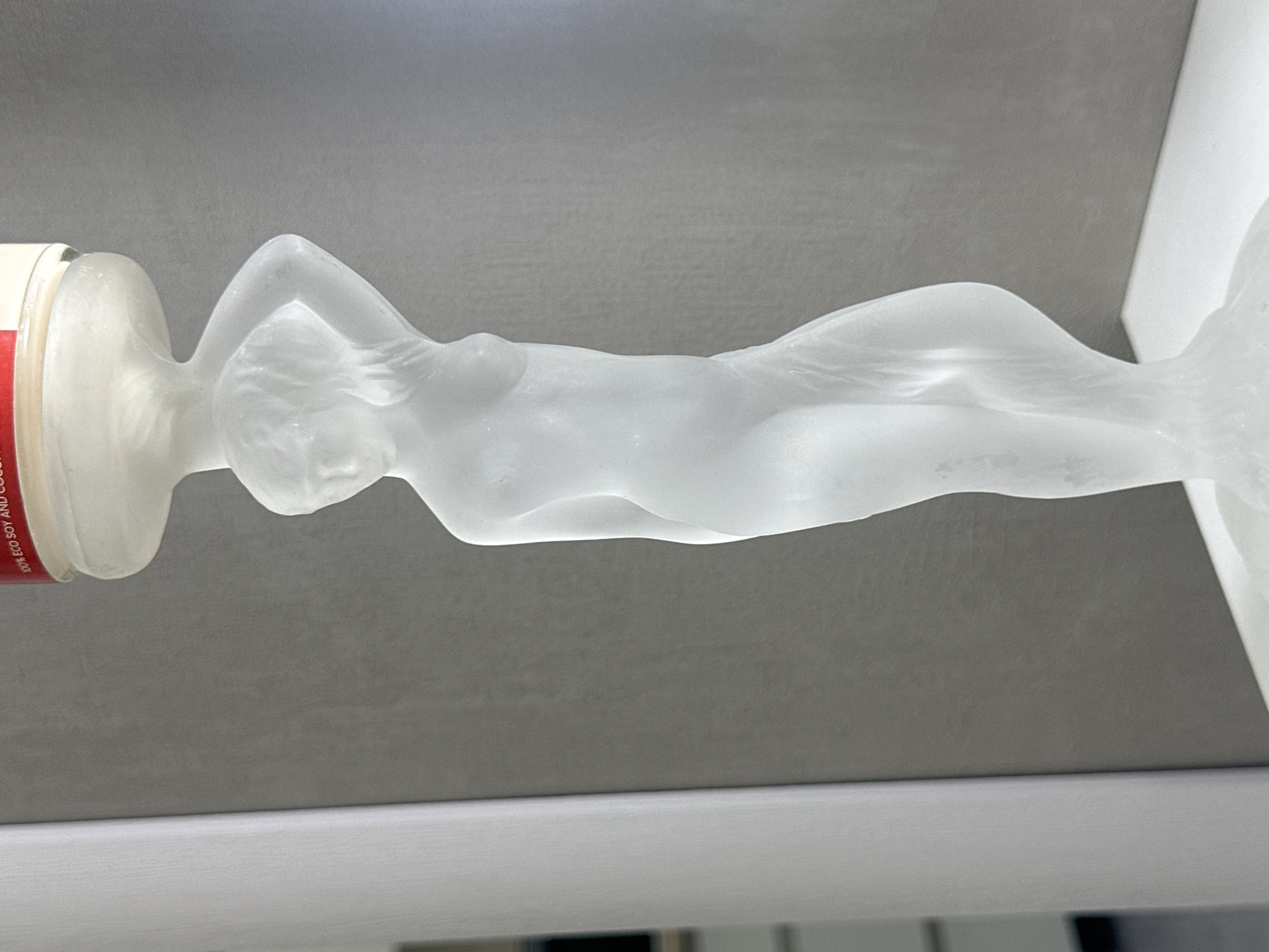 The image features a translucent, frosted glass sculpture of a reclining nude female figure. The sculpture appears to be resting on a white surface. A portion of a cylindrical object, possibly a container, is visible on the left with red and white text that mentions "% ECO SOY AND COCO". The background is a uniform light gray. There are no other objects, people, or locations visible to make an accurate decision to the city of Sochi, Russia.