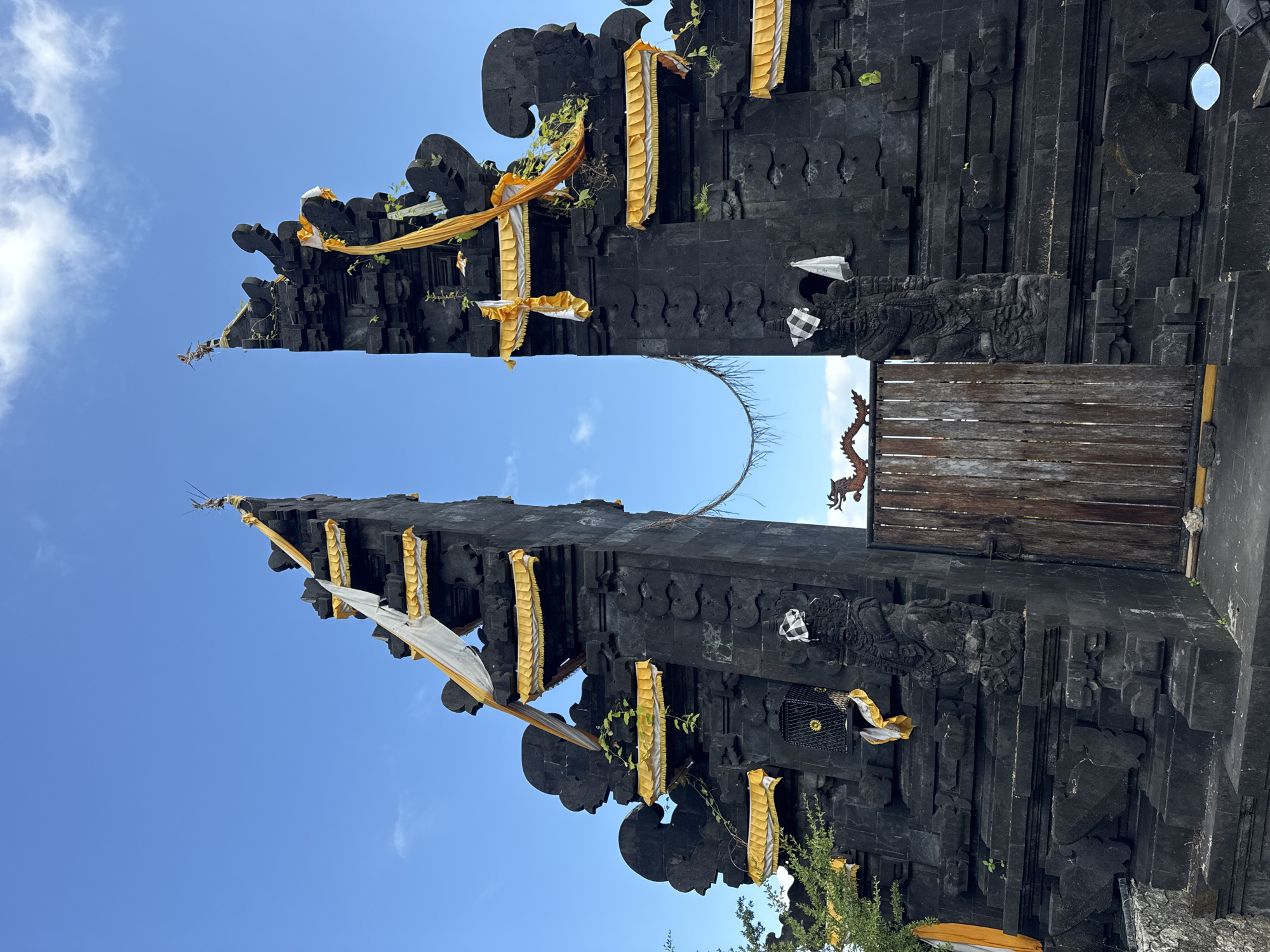 This media file captures a majestic, traditional Balinese temple gate, known as a Candi Bentar, crafted from dark volcanic stone. The gate is comprised of two tall, symmetrical halves that stand apart, creating an entrance in the middle.
The intricate stonework features multiple tiers, elaborate carvings, and decorative motifs characteristic of Balinese Hindu architecture. Adorning the structure are numerous pieces of yellow and white fabric, possibly ceremonial decorations or offerings, draped over the stone elements. Small patches of green foliage and vines are seen growing organically on the dark stone surfaces.
In the central opening, a rustic wooden gate, constructed from horizontal planks, appears to be closed. Perched atop the wooden gate is a decorative, reddish-brown figure that resembles a mythical creature or dragon. A slender, curved, bare branch or vine extends horizontally across the open space above the wooden gate, visually connecting the two stone gate halves.
The scene is bathed in bright daylight under a clear, vibrant blue sky, with only a few sparse, wispy white clouds scattered across. This suggests a sunny day, likely mid-day. The setting is outdoors, strongly indicative of a sacred site or temple entrance, consistent with the provided location context of Kuta Selatan, Indonesia. No people or specific activities are visible, emphasizing the grandeur and cultural significance of the architectural subject. There is no visible text in the image.