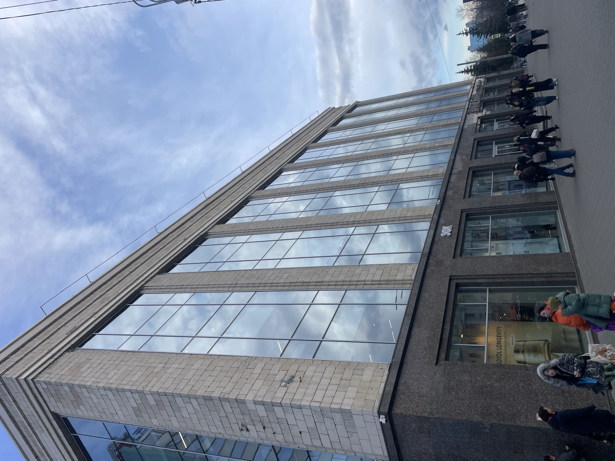 The image captures a vibrant urban street scene from a low-angle perspective, showing a large building and numerous pedestrians. The main subject is a multi-story modern building with a striking facade. The upper sections feature a light-colored, possibly stone or concrete, exterior with horizontal bands, contrasting with expansive glass windows that reflect the bright, partly cloudy sky. The lower floors of the building are darker, appearing to be made of granite or similar dark stone, with large display windows on the ground level.
Many pedestrians are walking on the sidewalk to the right of the building, moving in various directions. They are dressed in warm winter or cold-weather clothing, including thick coats, hats, and scarves, suggesting cool temperatures. One person in a bright orange jacket and green coat is visible closer to the foreground, while others appear further down the street near what looks like a pedestrian crossing.
The sky is a prominent feature, displaying a mix of light blue and scattered white, wispy clouds, which are beautifully mirrored in the glass windows of the building. The time of day appears to be daytime, with ample natural light. Overhead, power lines or street light wires are faintly visible against the sky. In the background, some bare trees and other city buildings are present, indicating a dense urban environment.
Visible text on the dark lower section of the building includes "БИЗНЕС-ЦЕНТР" (BUSINESS-CENTER) above an entrance. Inside a display window, promotional text for beauty products is visible, including "IDIOL LONGEVITY" and "VIHT," along with some Russian text. The overall scene depicts typical daily life in a bustling city.