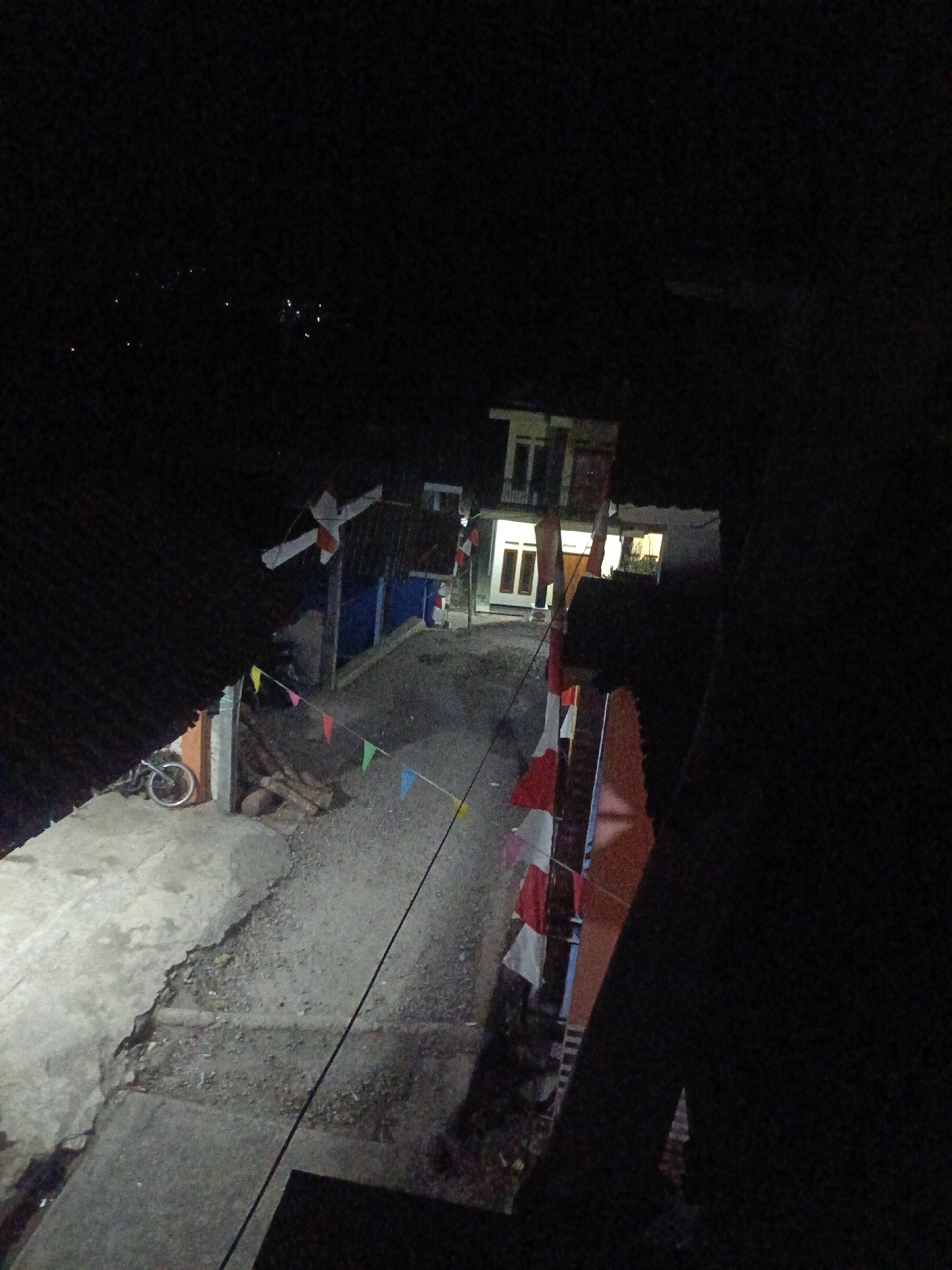 The image depicts a dark, possibly nighttime scene in Jambu, Indonesia, with some artificial lighting illuminating a residential area. In the foreground, on the left, there is a concrete surface next to a dark, corrugated roof structure. A bicycle is parked on the concrete. A string of colorful triangular flags stretches across a gravel path leading towards buildings in the background.
The background features a multi-story building with illuminated windows and doors. Red and white flags, possibly Indonesian flags, are displayed on poles and strings, suggesting a festive or patriotic occasion. The overall environment appears to be a narrow street or alleyway between houses. The lighting is dim, with shadows cast by the structures and the low ambient light. Distant lights are visible, possibly indicating other houses or a town in the distance. The ground is a mixture of concrete and gravel, with some scattered debris. The image quality is low, with significant noise and grain due to the low light conditions.