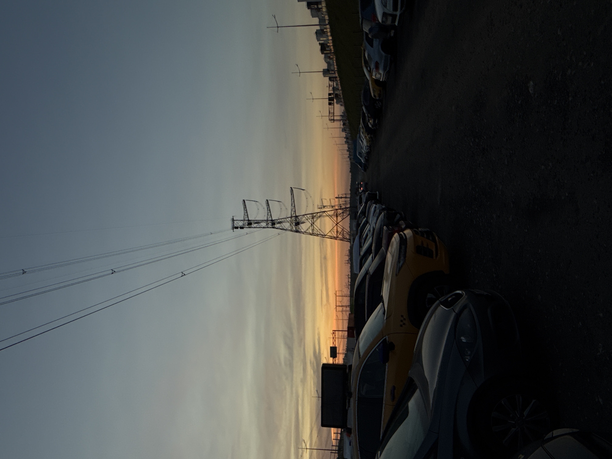 A dimly lit, wide-angle outdoor photograph captures a parking lot filled with cars at dusk. A tall, lattice-like power pylon dominates the right side of the frame, with its wires stretching diagonally across the sky. The sky transitions from a pale blue at the top to warm orange and yellow hues near the horizon, indicating the setting sun. In the foreground, several cars are parked, with their shapes and details obscured by shadows. A yellow taxi with a black sign on its roof is prominently featured on the left side. In the background, more cars are lined up, and a row of industrial-looking buildings or tanks can be seen on a gentle slope. The ground is a dark, gravelly surface. No people are visible in the scene. The location is identified as Severnoye, Russia.