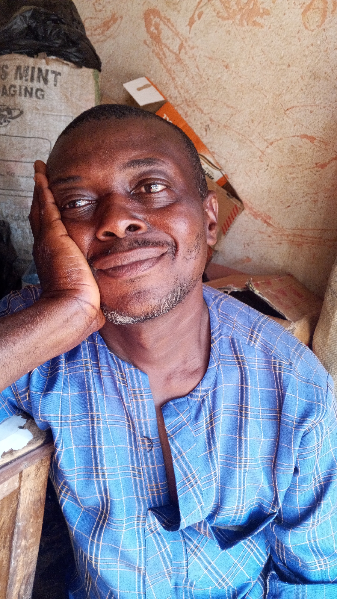 An adult male with brown skin and a patchy grey beard rests his head in his hand. He is wearing a blue plaid shirt with yellow stripes. Behind him are cardboard boxes, a burlap bag labeled "S Mint AGING" in block letters, and a black plastic bag. The background is a mottled beige wall with reddish-brown markings. The location is Sabon Tasha, Nigeria.