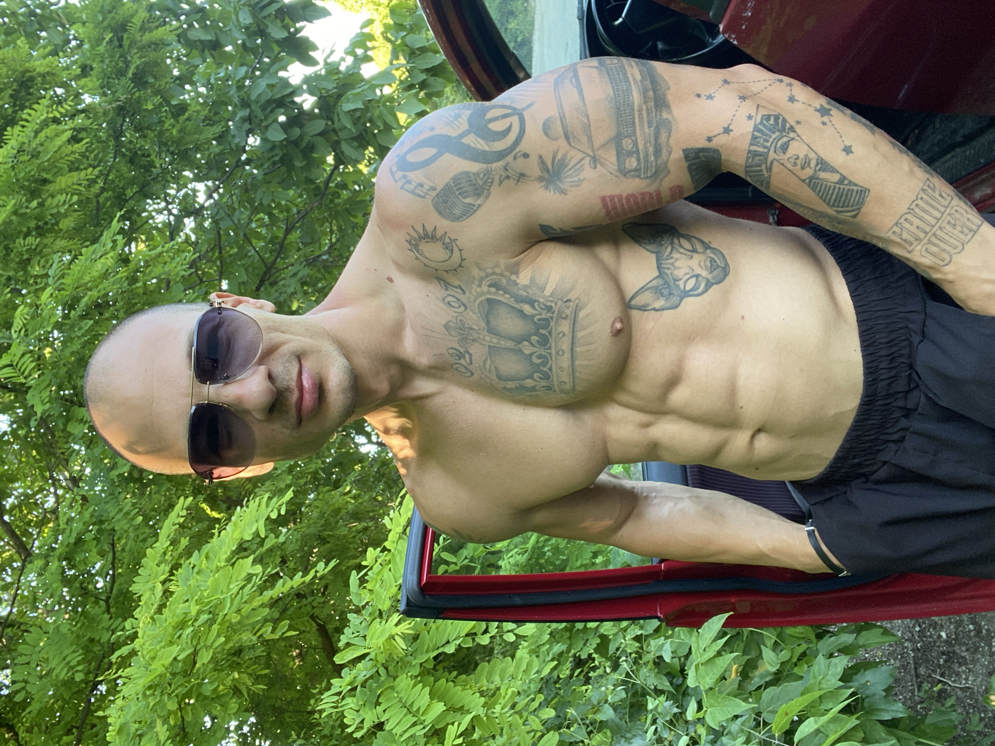 A man with a shaved head, wearing sunglasses and black shorts, stands next to a red car with the door open. He is shirtless, revealing multiple tattoos on his chest and arms, including a crown, a cat's face, musical notes, a car, and a pharaoh's head. The scene is set outdoors with lush green foliage in the background. The lighting suggests it is daytime, possibly late afternoon, with sunlight filtering through the leaves. The man's pose is relaxed, with his left hand in his pocket. There is no discernible urban environment visible, and no indicators suggesting the location is Pleven, Bulgaria.