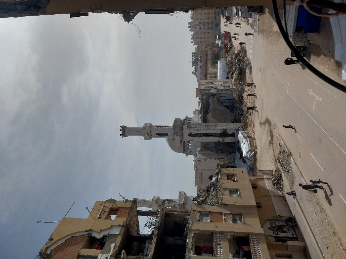 This image captures a wide street scene in Khan Yunis, Palestinian Territory, under a clear, bright sky with some atmospheric haze. The primary focus is a street flanked by heavily damaged buildings and substantial debris.
On the left, a multi-story building exhibits extensive structural damage, with several floors partially collapsed, exposing interior rooms, rebar, and broken masonry. Balconies with clotheslines are visible on some remaining sections. On the far right, a partial view of another damaged building frames the scene.
In the mid-ground, a wide asphalt road, marked with faded white lane lines and a directional arrow, is partially covered by dirt and scattered debris. Approximately 15-20 individuals are dispersed across the road and adjacent dirt areas, walking, some in small groups, others individually. Shadows indicate a daytime setting.
The central feature is a large mosque, significantly damaged, located behind a substantial pile of rubble and two white tarp structures. Its prominent white minaret appears largely intact, while the main mosque structure, including its dome, shows severe destruction with large sections of walls collapsed.
Further down the street, beyond the mosque, more urban structures are visible, some appearing damaged, and two red vehicles, consistent with emergency or utility trucks, are present. A small blue kiosk is partially visible on the bottom right. The environment is characterized by widespread destruction and ongoing human activity amidst the ruins.