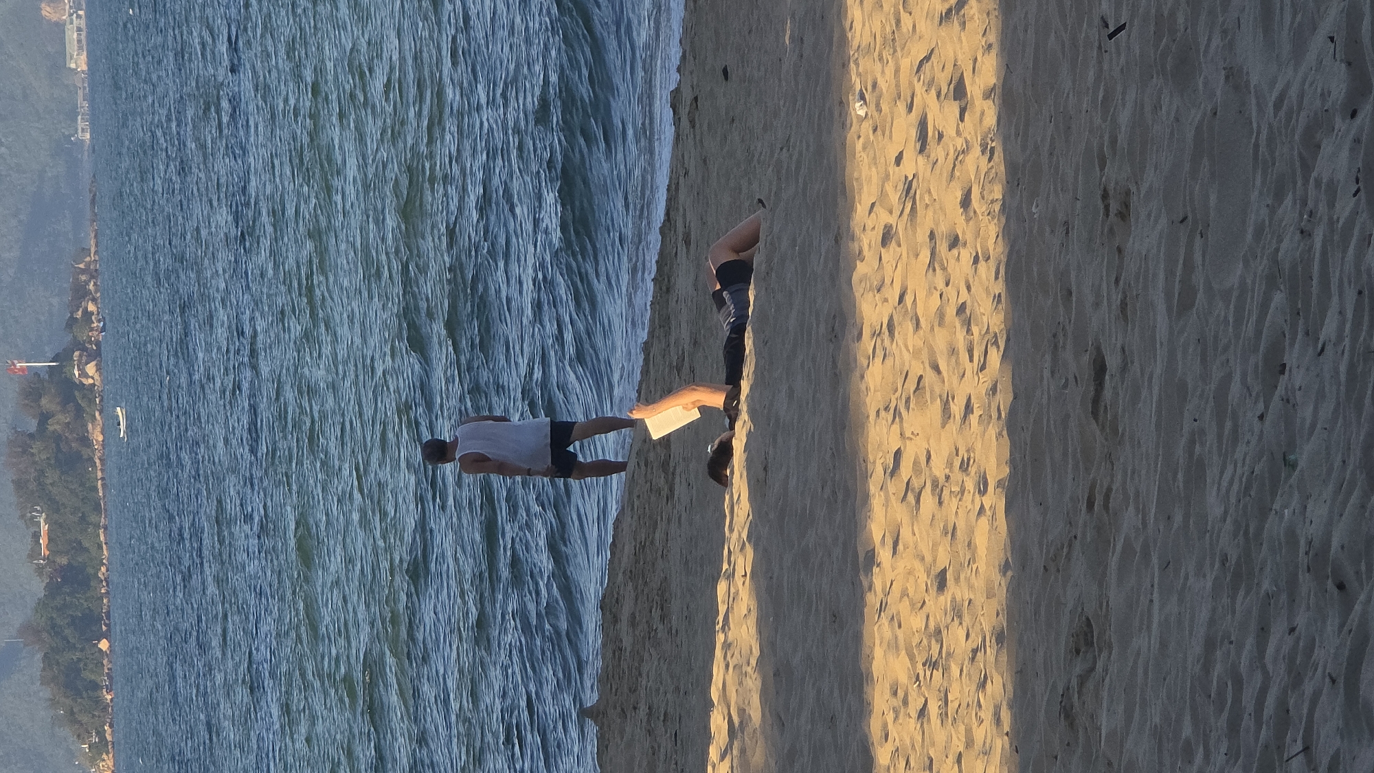 The image depicts a coastal scene at Hon Chong Promontory, Nha Trang, Vietnam, under natural daylight. The foreground consists of light tan sand, with a distinct shadow line horizontally bisecting the beach. In the sunlit portion of the sand, a person is lying supine, partially obscured, holding a white rectangular object (likely a book or tablet) vertically with their feet or lower legs.
In the mid-ground, a second individual stands in shallow ocean water, facing away from the viewer. This person wears a white tank top and dark shorts. The water exhibits moderate ripples.
The background features a body of water extending to a distant landmass identified as Hon Chong Promontory. This promontory is characterized by rocky formations and green vegetation. A prominent white flagpole flying a red Vietnamese flag is visible on the promontory. A small white boat floats offshore near the promontory. Further to the right, a low-lying structure, possibly a pier or building, extends over the water near the distant shore. A hazy mountainous or hilly landscape forms the horizon line.