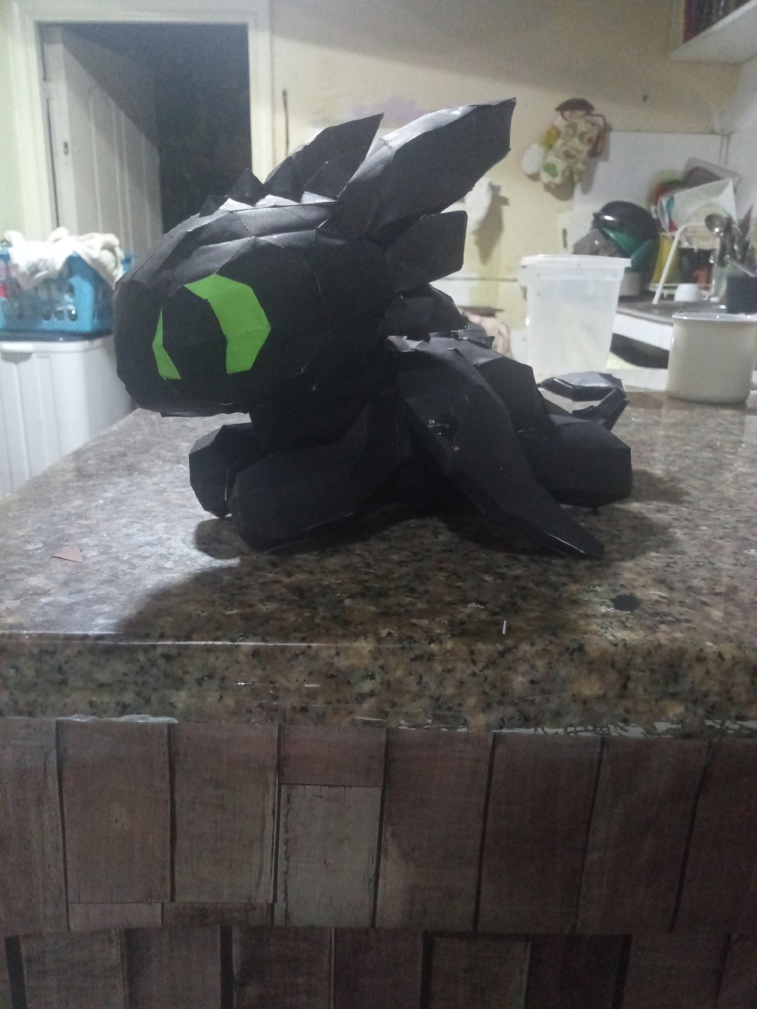 A low-angle, indoor shot shows a black paper-craft model of Toothless, the dragon from the movie "How to Train Your Dragon," sitting on a speckled, brown granite countertop. The model features green paper cutouts for eyes. The countertop is situated in a kitchen environment. In the background, a white door is partially open, revealing a dark space. To the left of the dragon is a blue laundry basket on top of a white dresser. In the right background, there is a collection of kitchen items including a clear container, white bowl, and miscellaneous utensils. The area below the countertop is constructed of vertical, dark brown wooden planks. The image does not provide any information that can identify the city of El Valle del Espíritu Santo, Venezuela.
