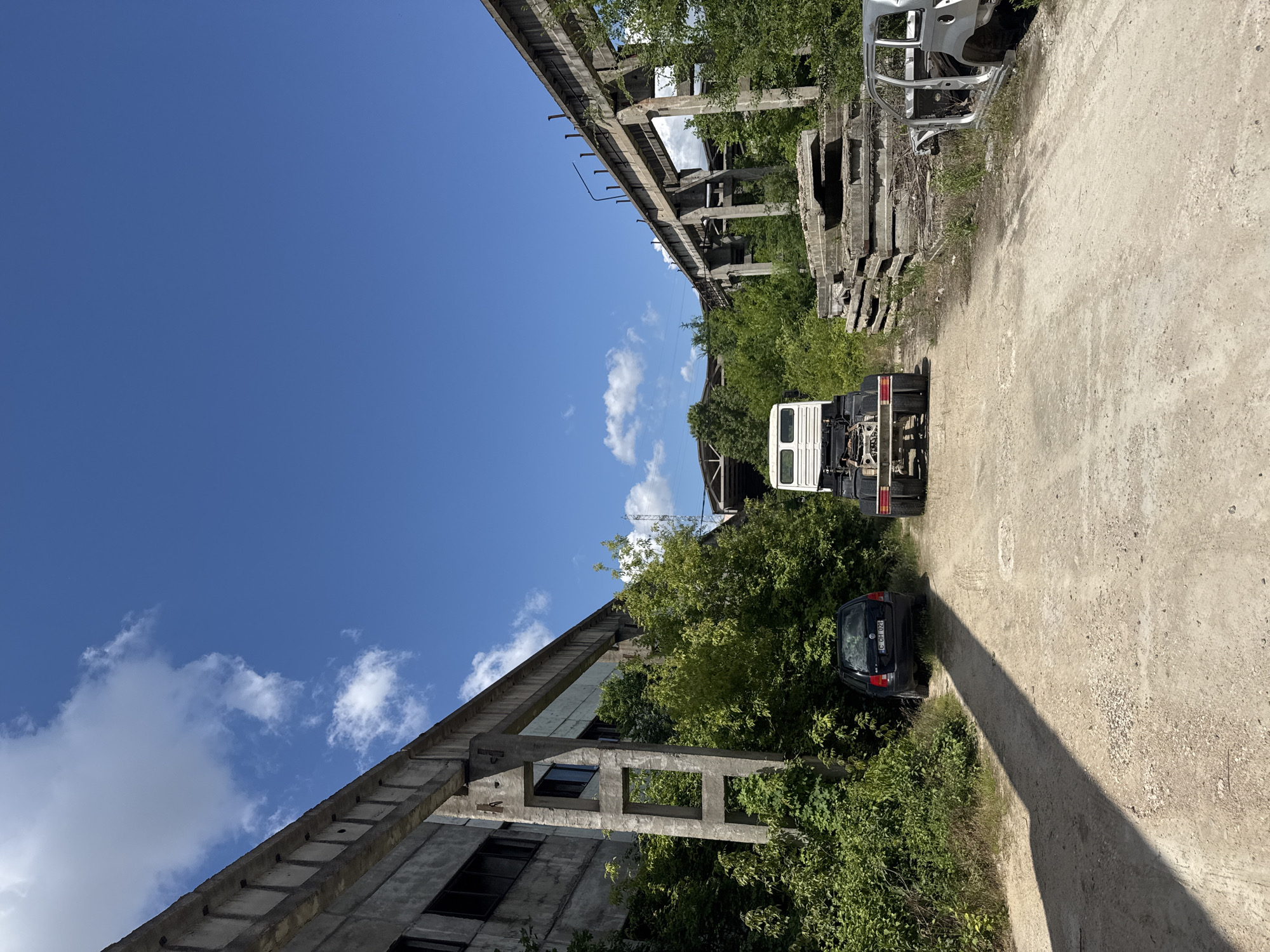 The image depicts an outdoor scene of an abandoned industrial area on a clear, sunny day. Two dilapidated concrete structures, likely remnants of old industrial buildings or overpasses, dominate the left and right sides of the frame. Lush green vegetation, including trees and bushes, has grown around and in between the structures, partially obscuring them.
In the foreground, a dirt path or road runs through the center of the scene. On the left side of the path, a dark-colored hatchback car is parked, partially covered by foliage. In the center of the path, a white truck chassis, without a cab or cargo bed, is positioned. To the right of the path, a stack of large, weathered concrete slabs is piled up, and further to the right, the stripped-down shell of a silver car is visible. The sky above is a vibrant blue with scattered white clouds. The overall impression is one of urban decay and nature reclaiming an industrial space.
