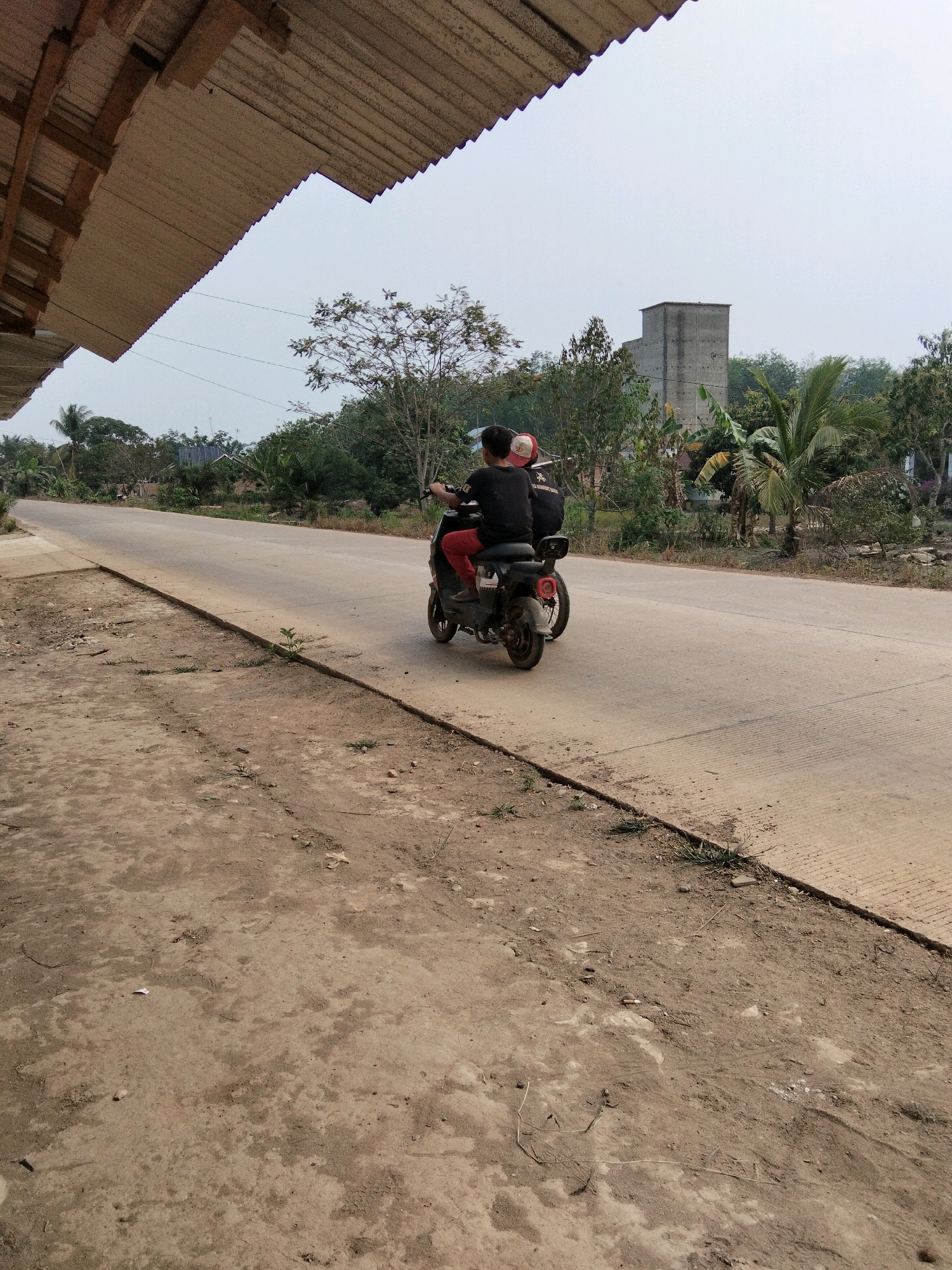 A motorcycle with two occupants travels on a paved road in Perambahan, Indonesia. The rider, wearing a black shirt, is positioned at the front, while a passenger in a black shirt and red pants sits behind. The motorcycle is dark-colored and has a rear cargo box. The road is flanked by a dirt shoulder on the left and vegetation and buildings on the right. A grey, multi-story building is visible in the background. The sky is overcast.