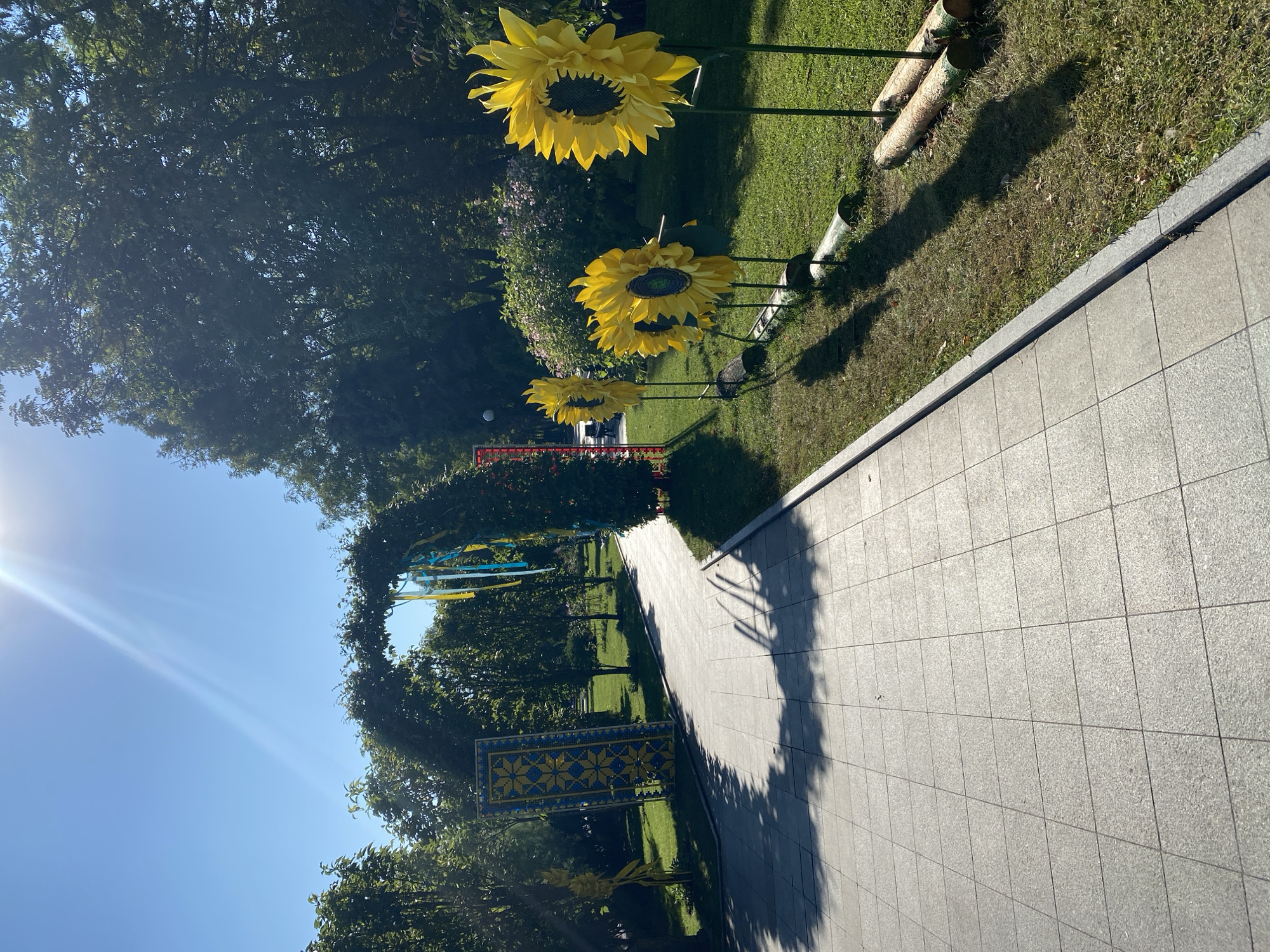 A sunlit park in Kremenchuk, Ukraine, features a paved walkway leading towards a green archway adorned with yellow and blue ribbons. To the right of the walkway, a row of large, artificial sunflowers stands on green poles, supported by logs at their bases. Beyond the sunflowers, a lush green lawn stretches towards trees and bushes. A decorative, tall rectangular panel with a yellow and blue geometric pattern is visible to the left of the walkway, near the archway. The sky is clear blue with the sun's rays creating a bright glare.