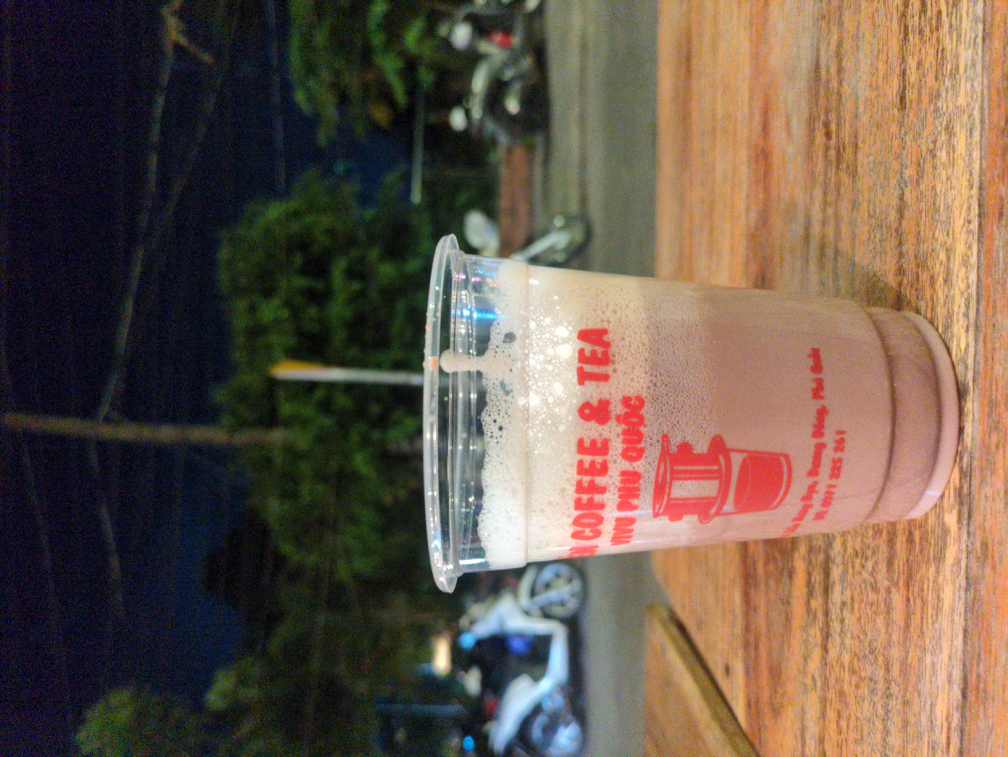 A plastic cup of iced coffee or tea rests on a wooden surface. The cup is translucent with a clear lid and the printed text "COFFEE & TEA" followed by "Phú Quốc" in a smaller font. A red logo resembling a traditional Vietnamese coffee filter is also visible on the cup. The liquid inside the cup appears to be a light brown color topped with a foamy layer.
The background is blurred, but suggests an outdoor setting at night. Visible elements include streetlights, trees, and parked motorbikes. The location is the city of Phú Quốc, Vietnam, as indicated by the cup's text.