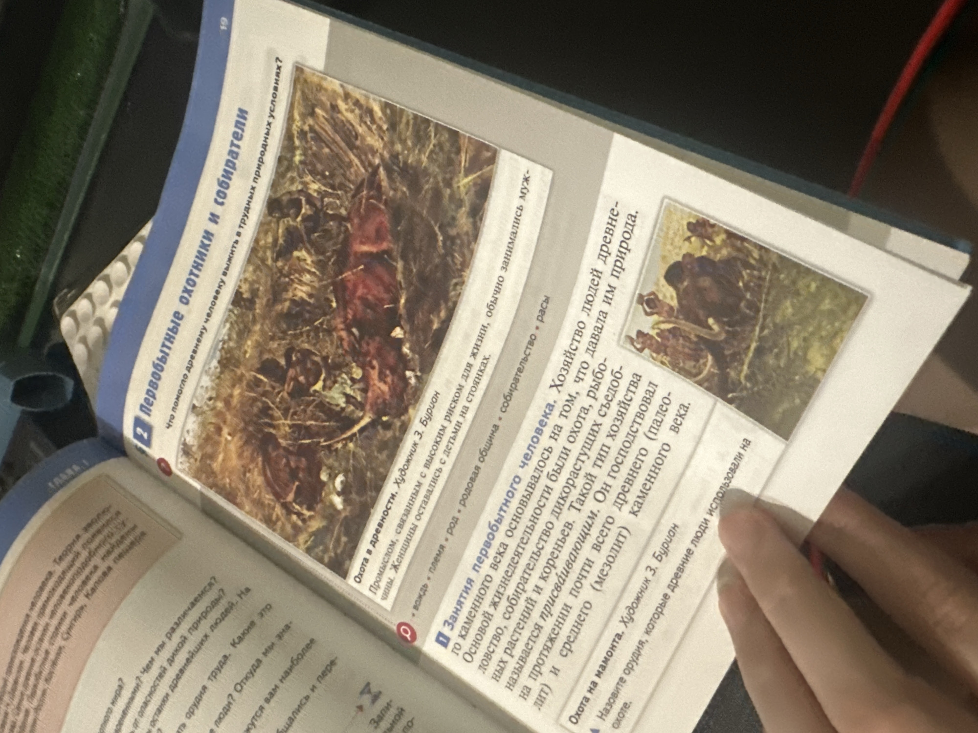 The image shows an open textbook on a table. The book contains text and illustrations about early humans, focusing on hunting and gathering. A hand is visible in the lower right corner, holding the page open. The lighting is soft and ambient.