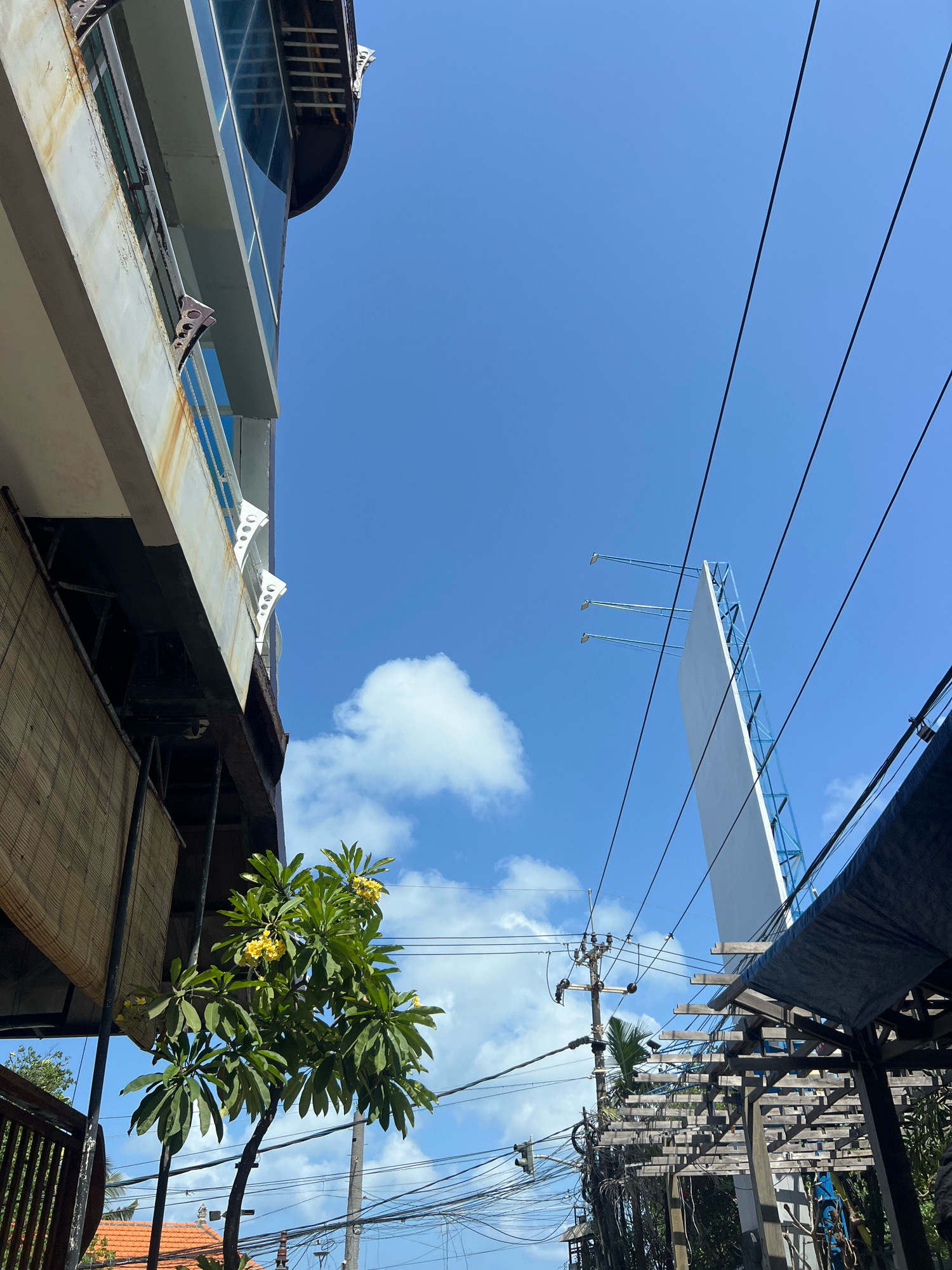 An exterior, low-angle, full shot captures a bright, sunny day in Kuta, Indonesia. The scene is dominated by a vivid blue sky with scattered white clouds. On the left, a portion of a modern, multi-story building is visible, featuring a weathered exterior with streaks of rust on its pale facade and large windows. Below this building, a bamboo blind covers part of the structure. To the right, a large, blank white billboard with a blue metal frame stands tall against the sky, intersected by a series of dark electrical wires. Further to the right, the wooden framework of an outdoor seating area with a dark blue canopy is partially visible. In the foreground, a lush green tree with yellow flowers is prominent, its branches reaching towards the sky. The presence of numerous electrical poles and tangled wires indicates an urban or semi-urban environment.