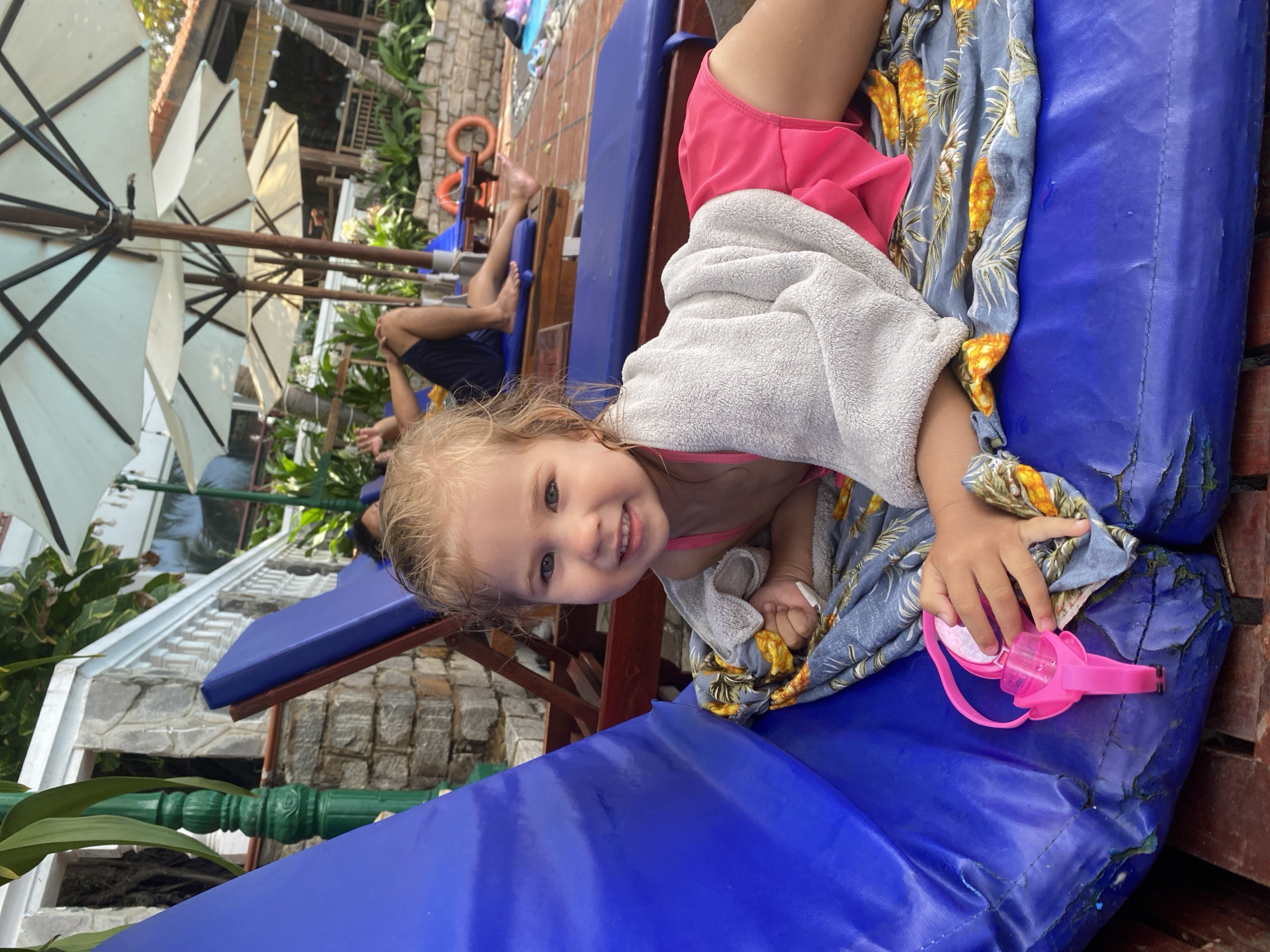 In this vertical, medium shot, a young girl with blonde hair is lying on a blue lounge chair near a swimming pool in Phu Thuy Ward, Vietnam. She is wearing pink shorts and a pink bathing suit, and is wrapped in a gray towel and a blue towel with a pineapple pattern. Her left hand holds a pair of pink swimming goggles. She is smiling at the camera, with a joyful expression.
In the background, several white umbrellas are set up, suggesting a sunny day. There are also stone walls and lush green foliage, characteristic of a tropical environment. Other people are visible in the background, some lounging on chairs and others walking. The scene appears to be a resort or a public pool area. The lighting suggests it is daytime, likely late morning or early afternoon. The overall mood is relaxed and cheerful, with the child's smile being the central focus.