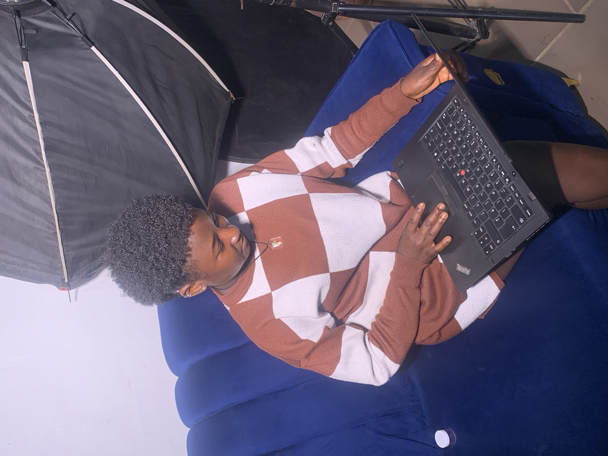 A young person with short, curly hair is lying down on a plush, blue couch, engrossed in using a black laptop. They are wearing a brown and white checkered sweater and a necklace. The setting appears to be indoors, possibly a studio, given the presence of photographic lighting equipment, including a large, black umbrella-like diffuser. The lighting suggests it might be daytime or artificial light is being used for a photoshoot. The person's expression is calm and focused, indicating they are concentrating on their work on the laptop. A small white container is visible on the couch near the person's feet. The laptop has "ThinkPad" branding visible on its surface.
