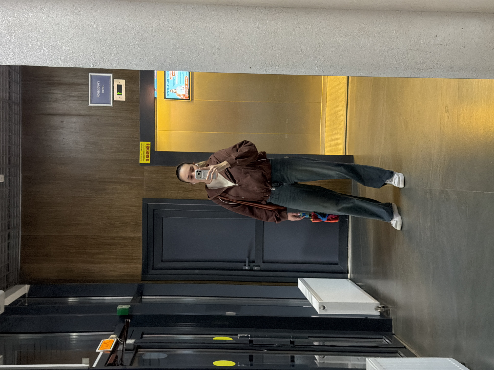 The image, taken indoors in a modern building hallway, shows a person taking a mirror selfie. The subject, a young adult with hair pulled back, is wearing a brown zip-up jacket over a light-colored top, dark jeans, and white sneakers. They hold a smartphone in their right hand to capture the reflection and a small, colorful object (possibly a toy or mini umbrella) in their left hand. Their expression is neutral.
The "mirror" is the highly reflective, gold-toned surface of closed elevator doors. To the left of the elevator doors, the wall is covered in dark wood paneling. Above a dark grey door, a blue sign with white Russian text is mounted. When correctly oriented and un-mirrored, the text reads "ТОЛЩ" (Tolshch) and "НОВОВОДНАЯ" (Novovodnaya). "Нововодная" is a street name, consistent with the given location context of Rostov-on-Don, Russia.
Adjacent to this sign, a small green digital display indicates "1", signifying the elevator's current floor. Reflected in the elevator doors is a yellow poster or screen, featuring mirrored Russian text that deciphers to "ИНВЕСТИРУЙ В ЯРКИЕ НОВЫЕ ИДЕИ!" ("INVEST IN BRIGHT NEW IDEAS!"). Below this reflection, a yellow sticker displays the number "7 829 825 000 8", likely a phone number.
The floor is dark grey tiled. A white radiator unit is partially visible in the bottom right foreground. The setting appears to be a well-lit indoor space, such as an apartment building or office lobby. The time of day and weather are not determinable from the artificial lighting.