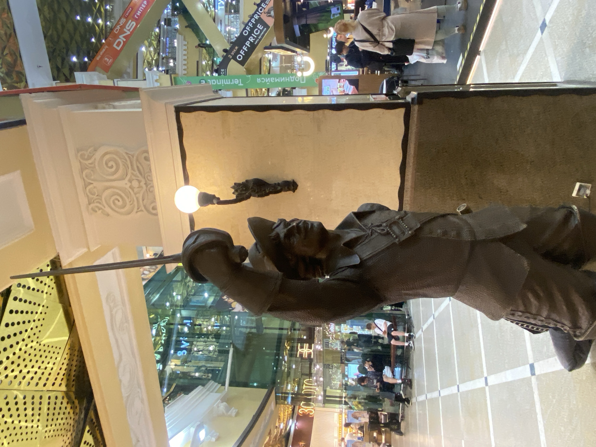 The image captures the interior of a large, multi-level shopping mall or commercial complex. In the foreground, positioned prominently, is a dark-colored statue of a man, likely a historical figure such as a musketeer or swordsman. He is depicted wearing a wide-brimmed hat, period clothing, and holds a long, thin sword upright in his right hand. A glowing wall lamp is mounted on the light-colored wall directly behind the statue.
The background reveals the bustling atmosphere of the mall, with several people visible on different levels. Some individuals are seen walking or standing, while others are seated on benches. The setting features modern architecture with glass railings and reflective surfaces, contributing to a bright, well-lit environment, presumably from artificial lighting.
Various commercial signs and storefronts are visible on upper levels. Notable text includes "DNS ГИПЕР" (DNS HYPER), likely indicating a large electronics retailer, and "OFFPRICE," a discount store chain, accompanied by the phrase "сеть аутлетов" (network of outlets) and "3 ЭТАЖ" (3rd FLOOR). Directional signage like "Поднимайся" (Go up / Ascend) is also present. Other visible fragments of text include "Terminal," "30 OTO," and "PERFI," suggesting a diverse range of businesses within the complex. The presence of Russian text confirms the location context of Yekaterinburg, Russia. The overall impression is that of a busy, contemporary urban shopping destination.