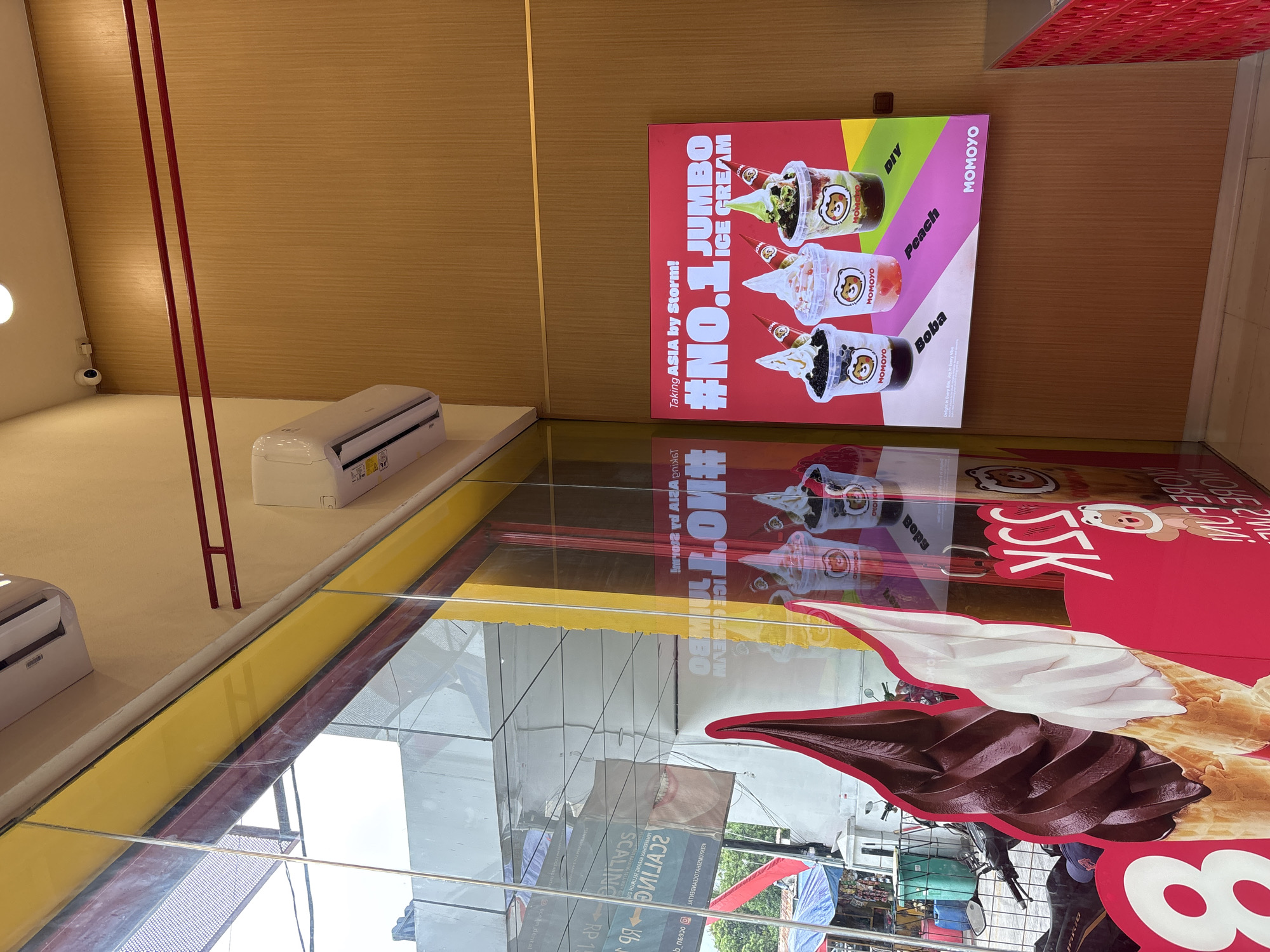The image shows the interior of a Momoyo ice cream shop, likely located in North Jakarta, Indonesia, based on the text and references on the signage. The shop features advertisements for "No.1 Jumbo Ice Cream" with flavor options like DIY, peach, and boba. Another large advertisement features a chocolate and vanilla swirl ice cream cone promotion for SSK. The reflection in the glass shows a bustling city street.