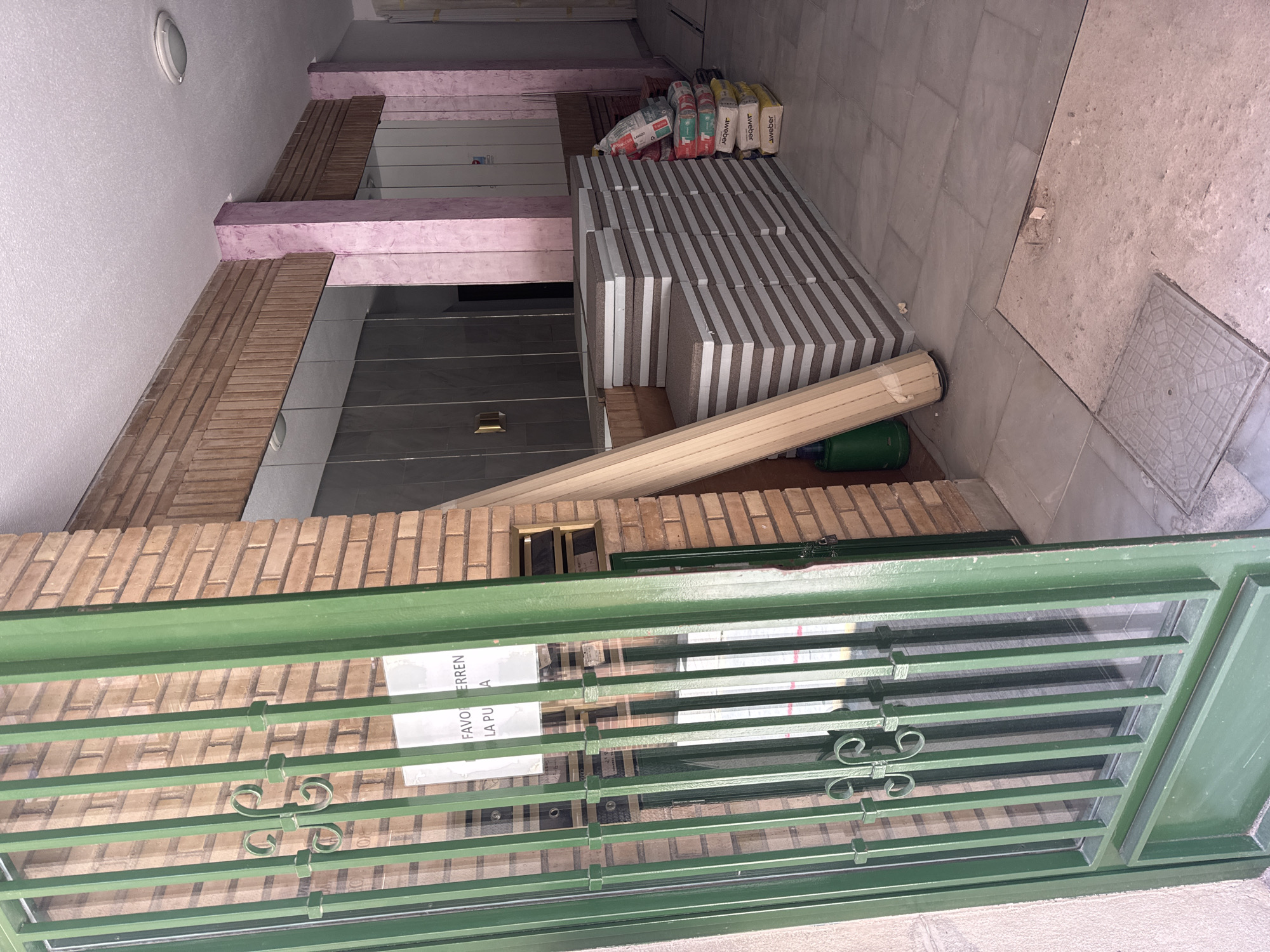 The image depicts a residential entryway under renovation. Piles of construction materials, including insulation panels and bags of mortar, are stacked on a tiled floor. A roll of bamboo matting leans against a wall constructed of brick. The entrance is partially obscured by a green metal grate with decorative scrollwork. A sign attached to the grate reads "FAVOR CERRAR LA PU..." which translates to "PLEASE CLOSE THE DOOR...". The scene suggests a domestic construction or repair project is in progress. The lighting is dim, suggesting it is either early morning or late afternoon, or possibly an indoor space with limited natural light. The location context of Murcia, Spain, is a useful detail for understanding the architectural style and materials used.