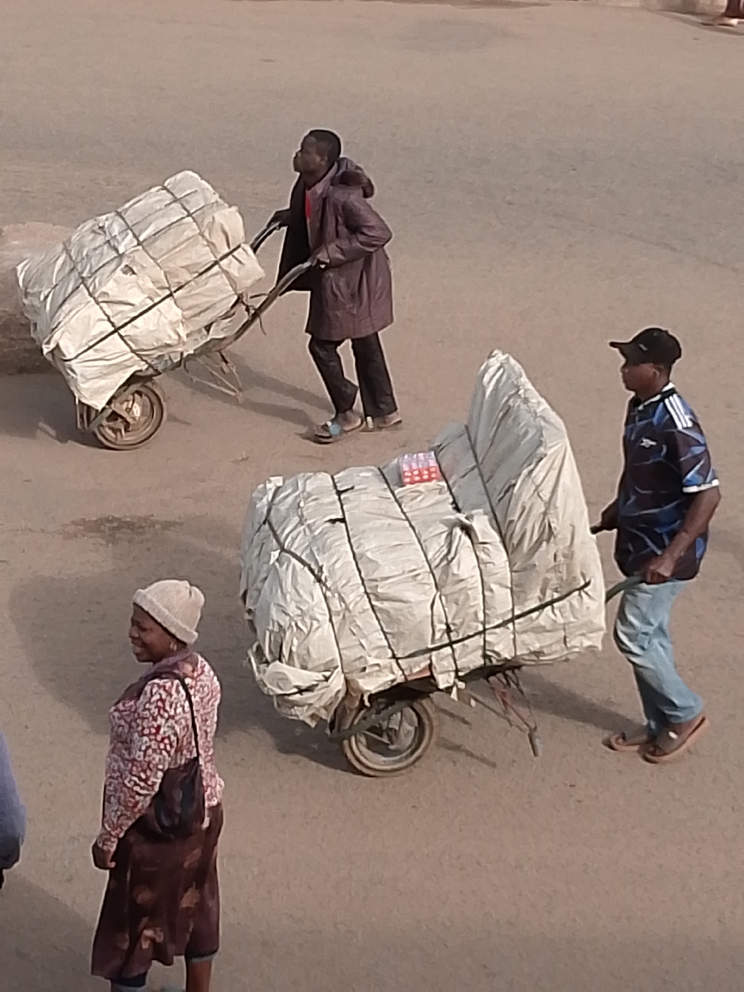 In Bukuru, Nigeria, three individuals are engaged in moving large, white-wrapped bundles on wheelbarrows across a dusty, paved surface, likely a street or market area. The scene is observed from a high angle.
On the left, a person wearing a dark coat and a red scarf is pushing a wheelbarrow laden with a substantial white bundle secured with black straps. This person appears to be walking away from the viewer.
In the center and slightly to the right, another person, a woman wearing a light-colored beanie, a patterned long-sleeved shirt, and a long skirt, is standing and looking to her left. She has a dark bag slung over her shoulder. She is positioned near the foreground.
To the right, a man wearing a black baseball cap, a blue and black patterned short-sleeved shirt, and light blue jeans is pushing a wheelbarrow. His wheelbarrow carries a similarly large, white-wrapped bundle, also secured with black straps. Visible on top of this bundle are some small, rectangular red and white packages, possibly food items. This man is walking towards the left of the frame, away from the viewer.
The ground is a dull, light brown, suggesting a dry, possibly arid environment. The lighting indicates it is daytime, with shadows cast by the people and the wheelbarrows. The overall impression is one of everyday labor and commerce in a Nigerian urban setting.