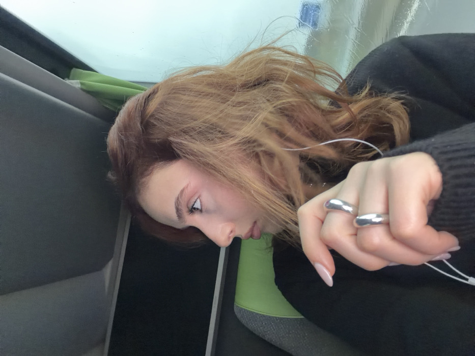 The image depicts a young adult female seated inside a vehicle, identified as being in Moineville, France. The subject is shown in profile, facing to the right of the frame, gazing upward and outward through a window. She has wavy, reddish-brown hair with lighter highlights. Her left hand is prominently featured in the foreground, wearing two large, smooth, silver-colored rings on her ring and middle fingers. Her fingernails are manicured with a light, neutral polish. White wired earbuds are visible, with the cord extending from her left ear. She is wearing a dark, possibly black, long-sleeved garment. To her left, part of a green seat or headrest is visible. To her right, a window displays a blurred, indistinct exterior, suggesting motion or an overcast environment, with visible moisture or streaks on the glass. A light green curtain or blind is partially visible at the top right edge of the window frame. The vehicle's interior ceiling, dark grey in color, is visible above her.