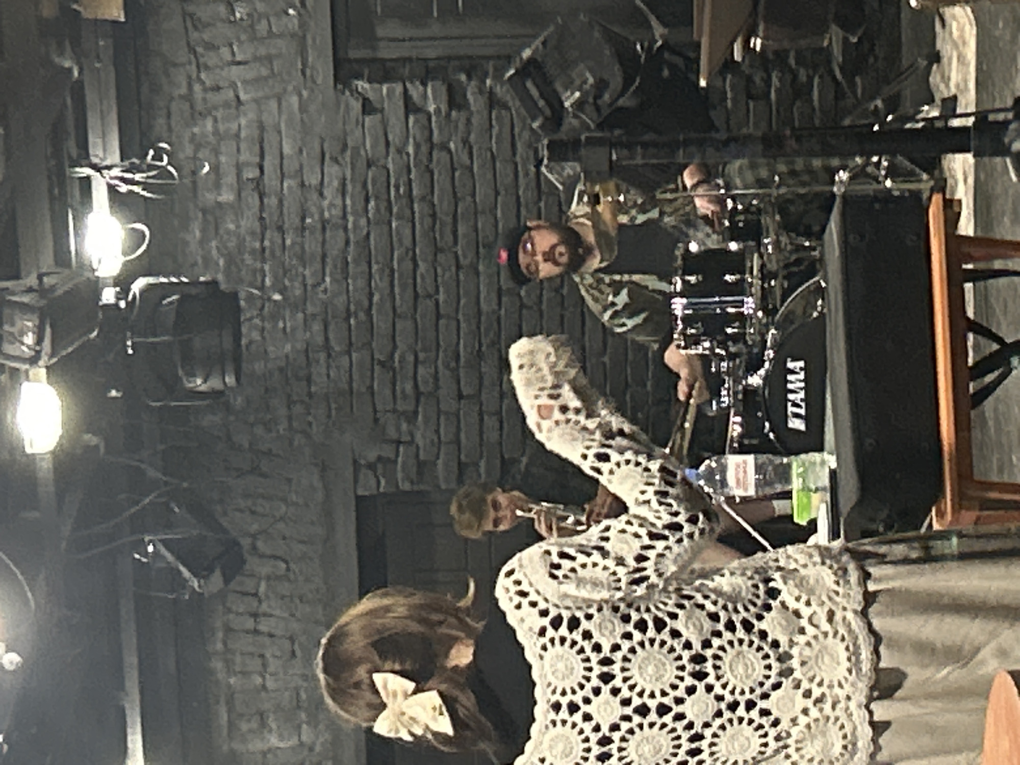 Here's a detailed description of the image:
**Scene Overview:** The image captures a live musical performance, possibly indoors, given the backdrop of what appears to be a brick wall.
**People:**
* A female figure is in the foreground, partially visible with brown hair adorned with a bow. She is wearing a white lace garment.
* A male drummer is playing behind the woman. He has a beard and is wearing a cap and patterned shirt.
* A male trumpet player is partially visible behind the woman, playing the trumpet.
**Objects:**
* A drum set, identifiable by the "TAMA" logo on the bass drum, is present.
* Stage lighting fixtures are visible near the ceiling.
* A bottle of water is near the drums.
**Actions:**
* The musicians are actively performing.
* The female figure is likely watching the performance, or perhaps part of it.
**Environment:**
* The performance takes place in a venue with exposed brick walls.
* The lighting is somewhat dim, with spotlights focused on the stage.
**Location:** The city and specific location are Unknown, Unknown.