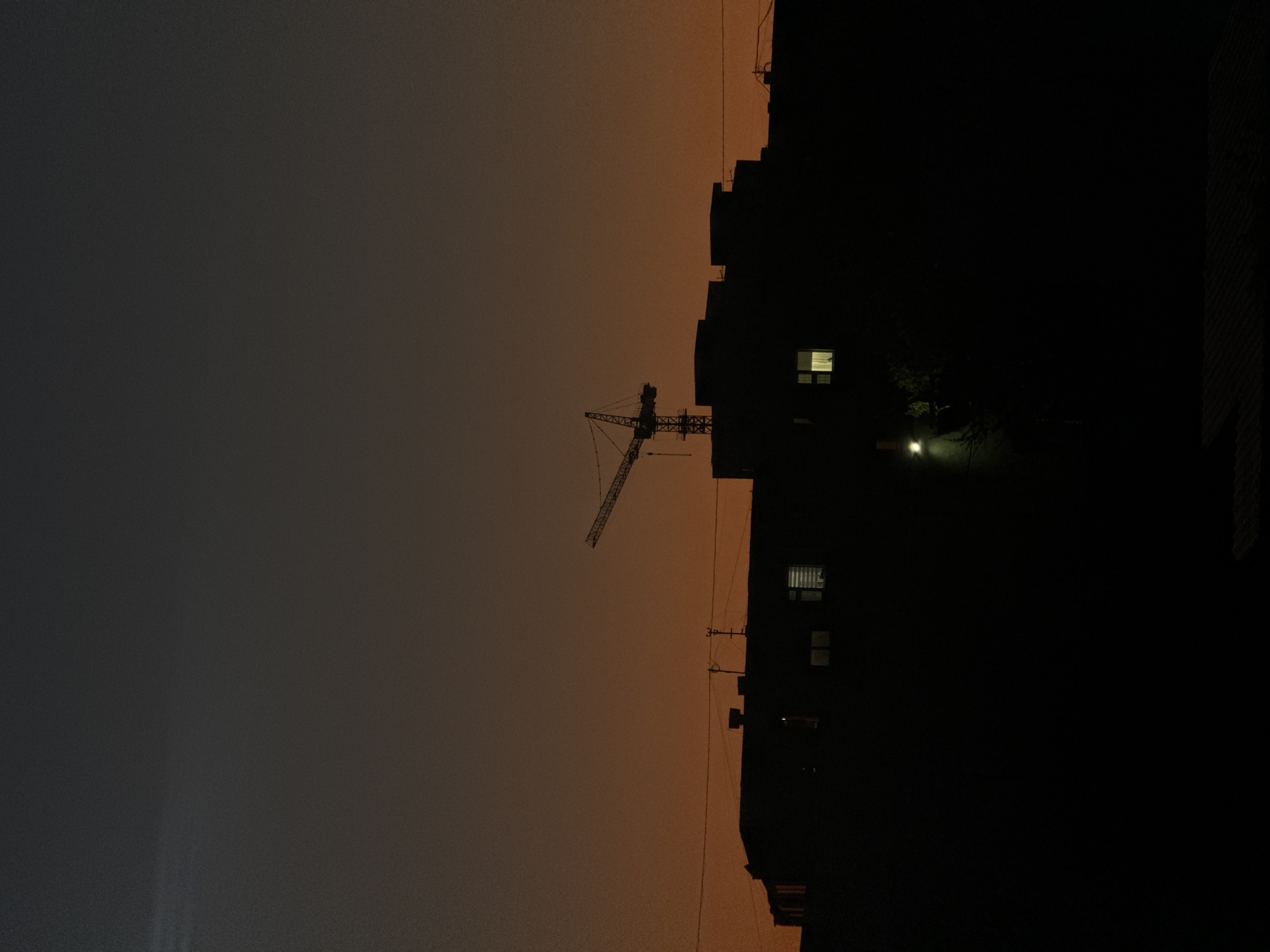 The image depicts a silhouetted cityscape at dusk, likely in Aktau, Kazakhstan. The sky transitions from a dark gray at the top to a warm orange near the horizon. Prominently featured in the mid-ground is a construction crane, its skeletal frame stark against the fading light. Below the crane, the dark silhouettes of several multi-story buildings stretch across the frame. Some windows in these buildings are illuminated, suggesting interior lights are on. A street light casts a faint glow on the lower left side of the scene, illuminating some trees. The overall atmosphere is quiet and still, with no people or active movement visible, emphasizing the architectural elements and the twilight sky.
