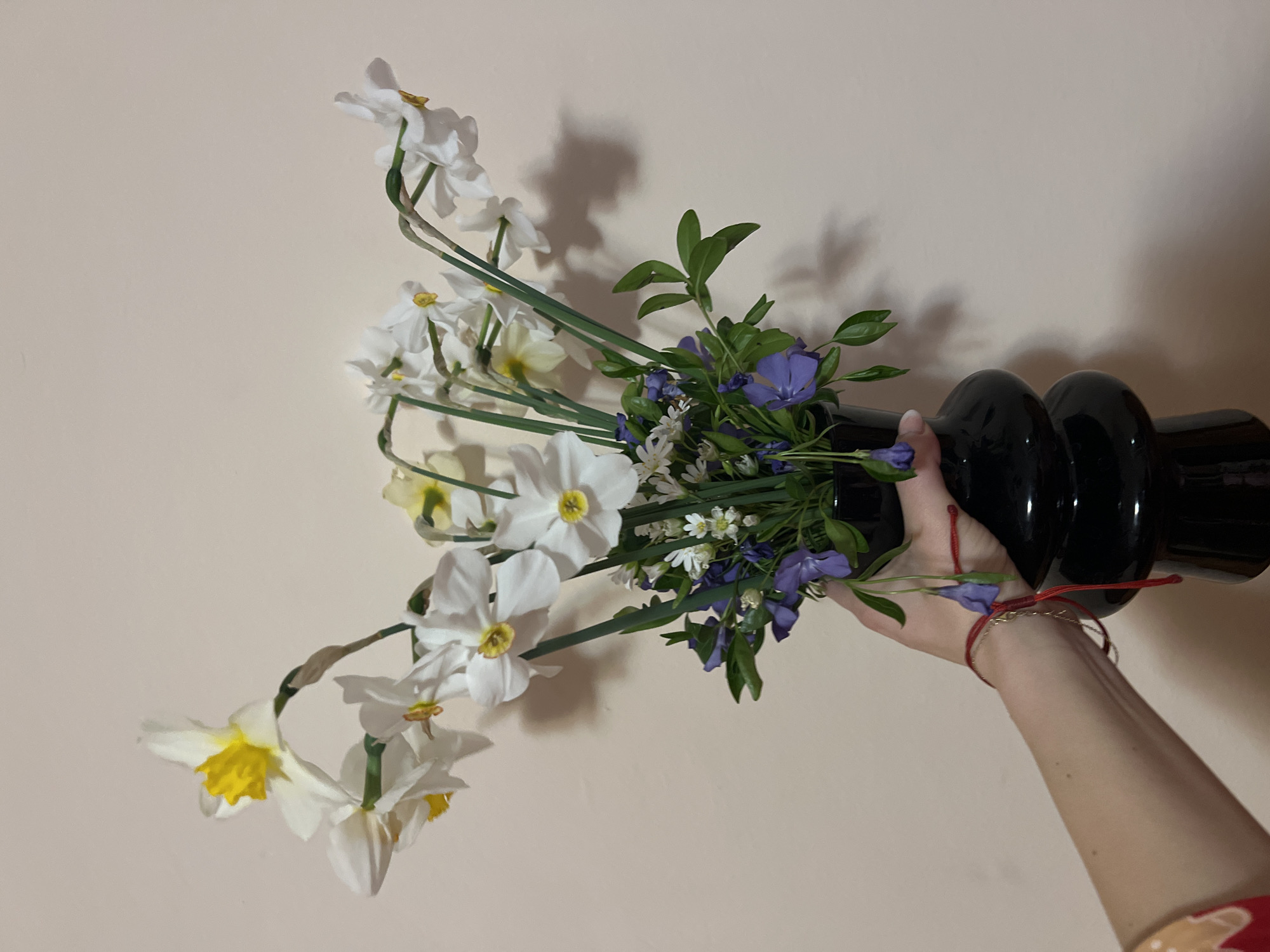 A hand holds a black vase filled with white daffodils, small blue and white flowers, and green foliage against a pale beige wall. The bracelets on the hand are visible. It is not possible to determine if this image was taken in Plovdiv, Bulgaria.